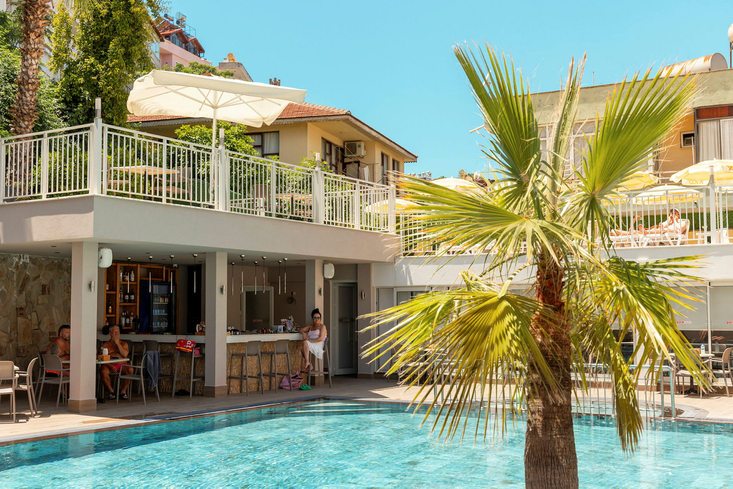 En swimmingpool med en bar intill och en palm i förgrunden. I bakgrunden syns en byggnad och en terrass med bord och parasoll. (AI-genererad text)