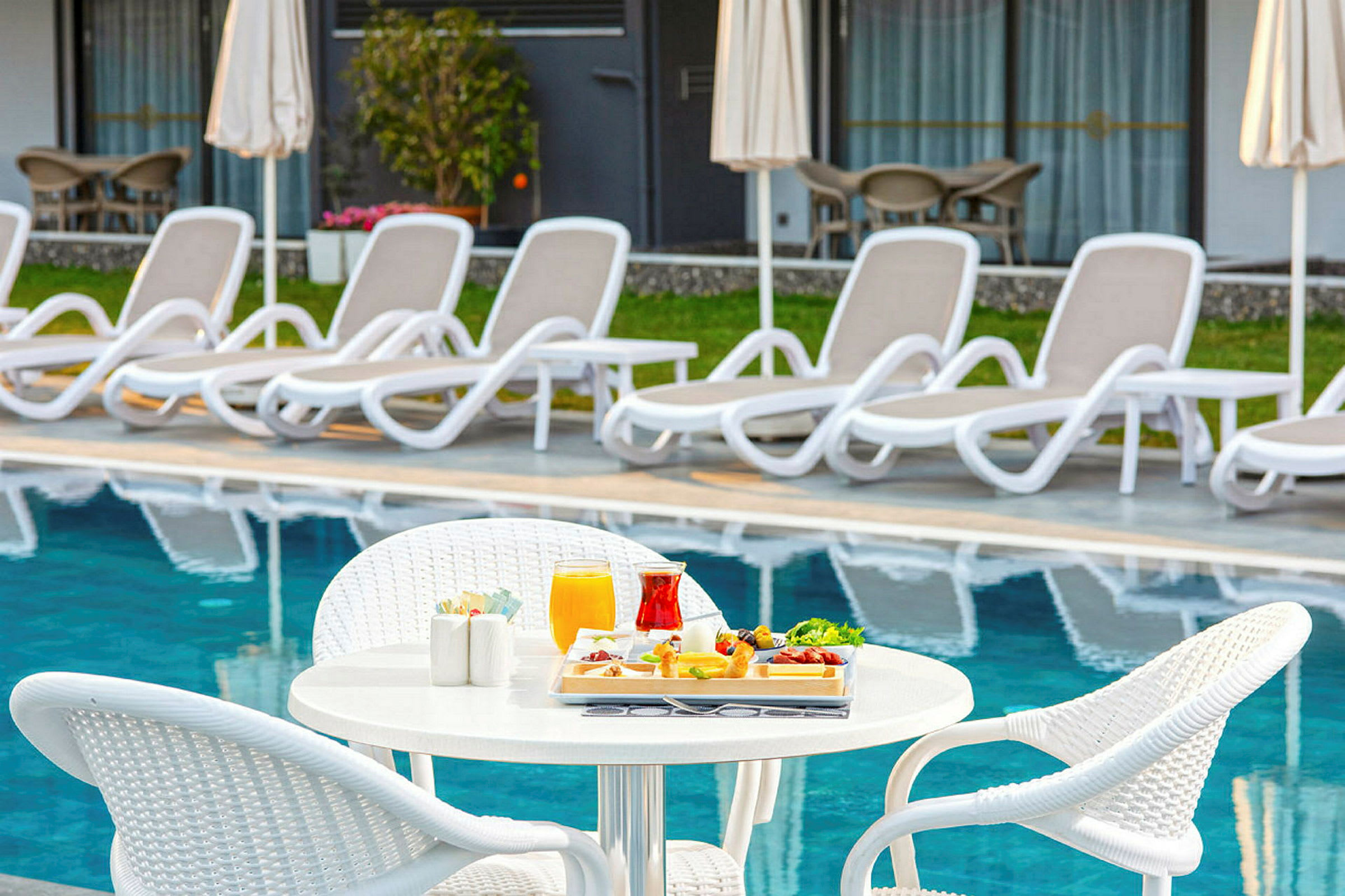 Ett bord med serverad mat och dryck intill en pool med solstolar i bakgrunden. (AI-genererad text)