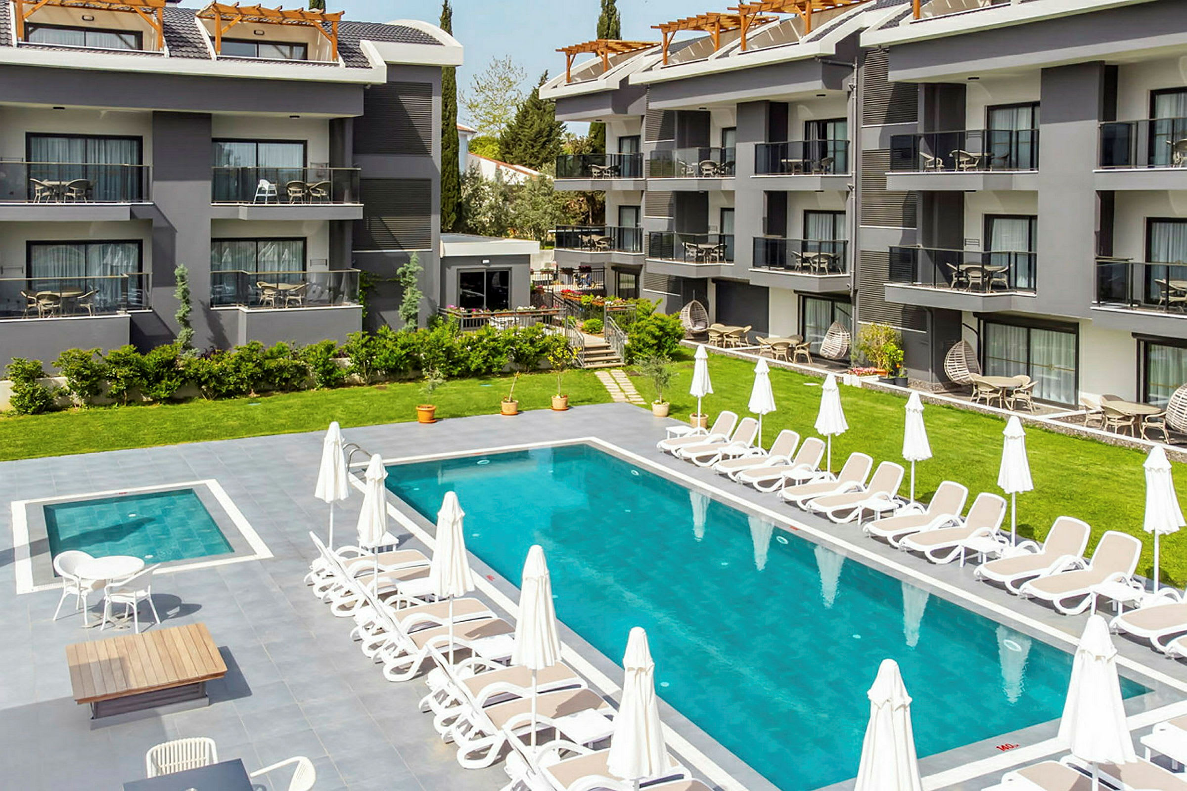 Ett hotellområde med en swimmingpool omgiven av solstolar och parasoller, samt byggnader med balkonger i bakgrunden. (AI-genererad text)