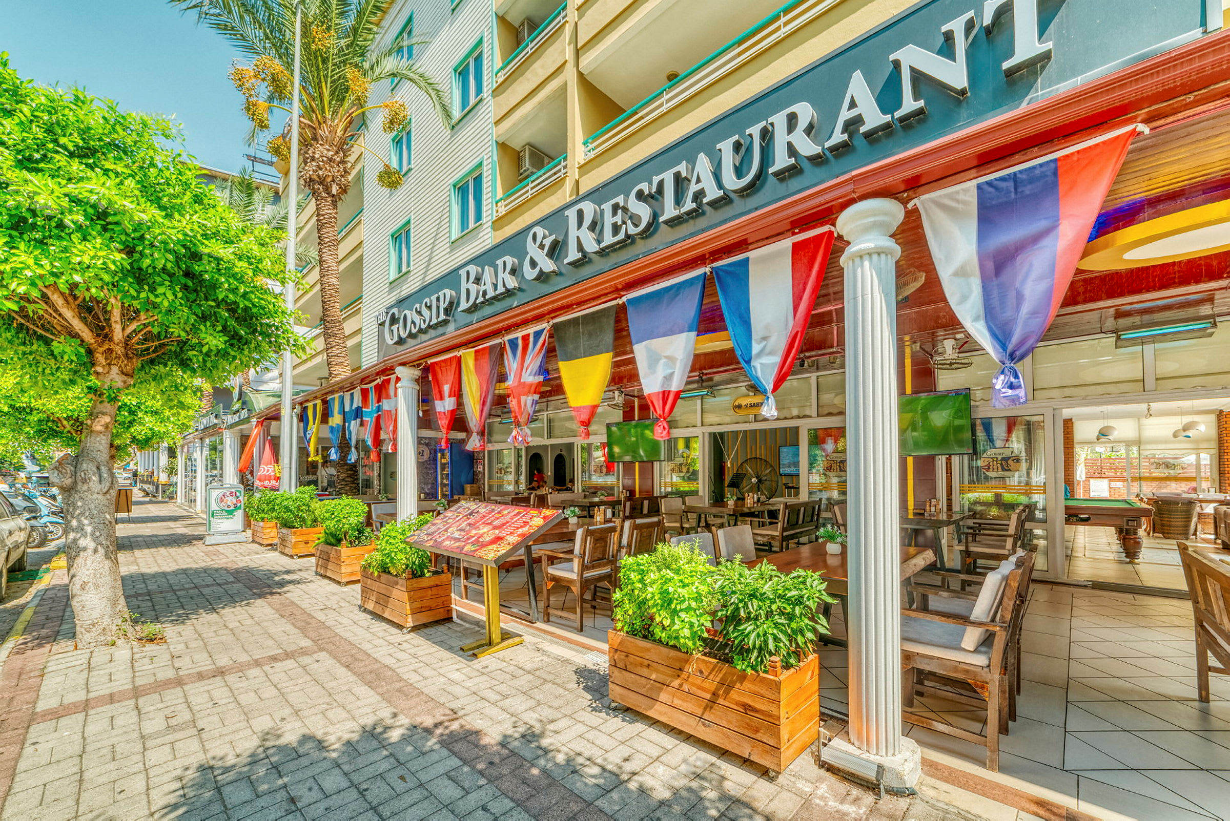 Ett café eller restaurang med uteservering, dekorerad med flaggor och omgiven av träd och grönska. (AI-genererad text)