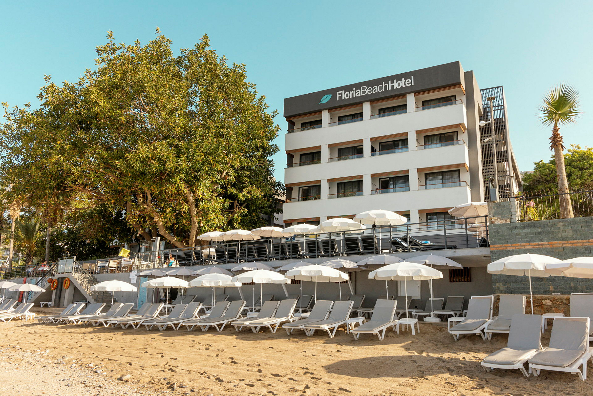 Vit byggnad med skylt 'Floria Beach Hotel' och en sandstrand med solstolar och parasoller. (AI-genererad text)