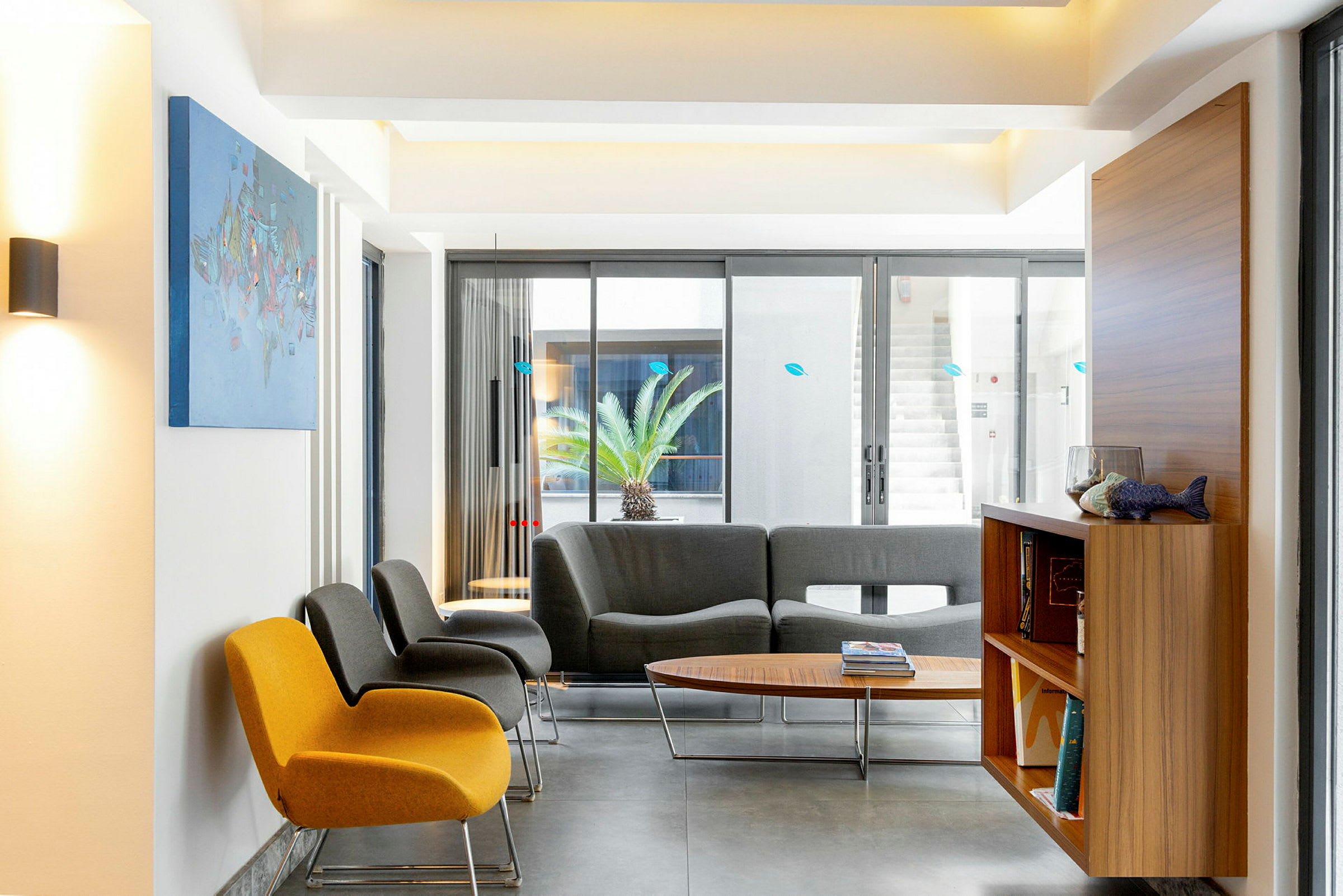 Ett modernt väntrum med stolar i olika färger och ett soffbord. Utanför syns en palm genom stora glasfönster. (AI-genererad text)