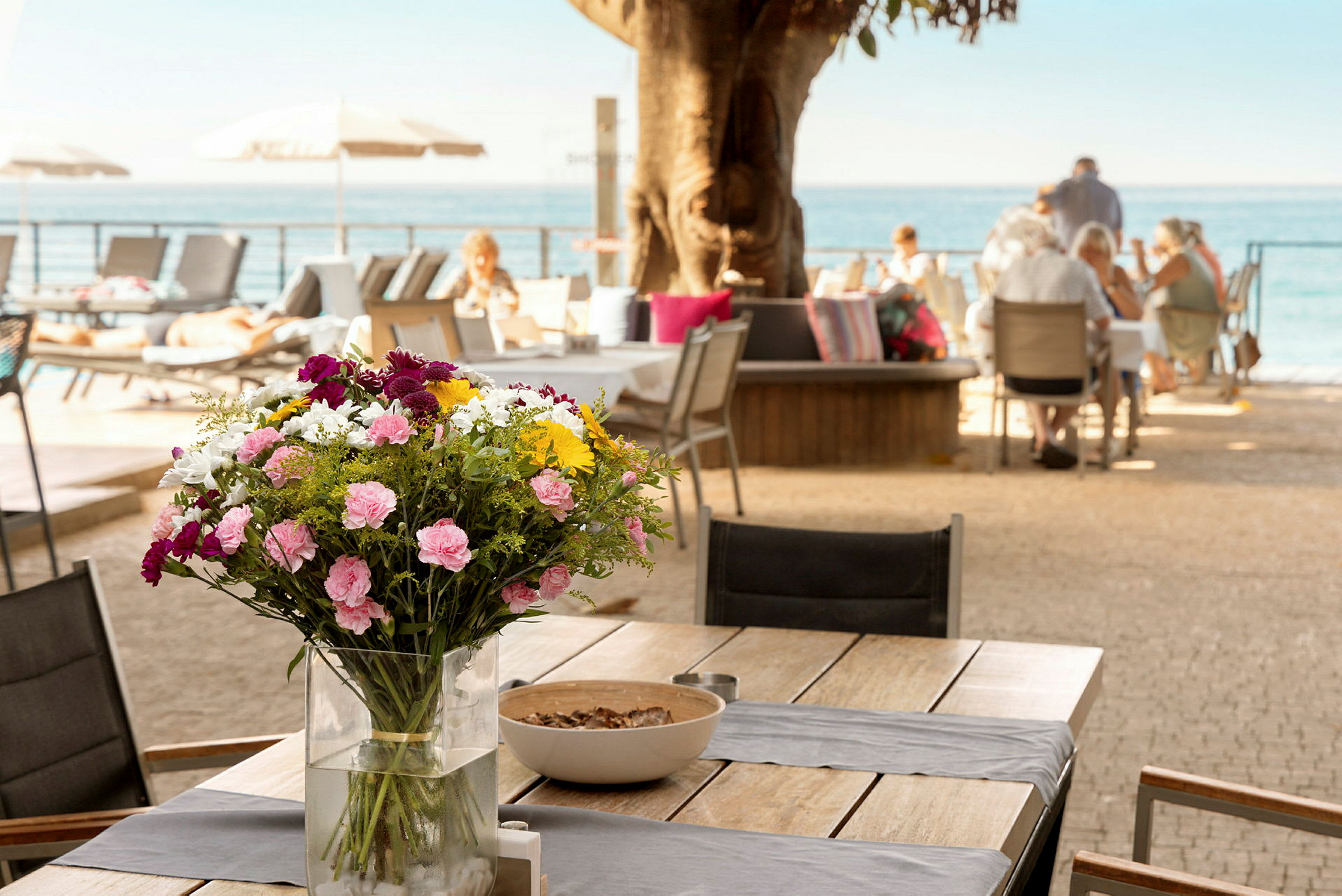Ett bord med en vas fylld med färgglada blommor, med människor som sitter vid andra bord i bakgrunden nära havet. (AI-genererad text)