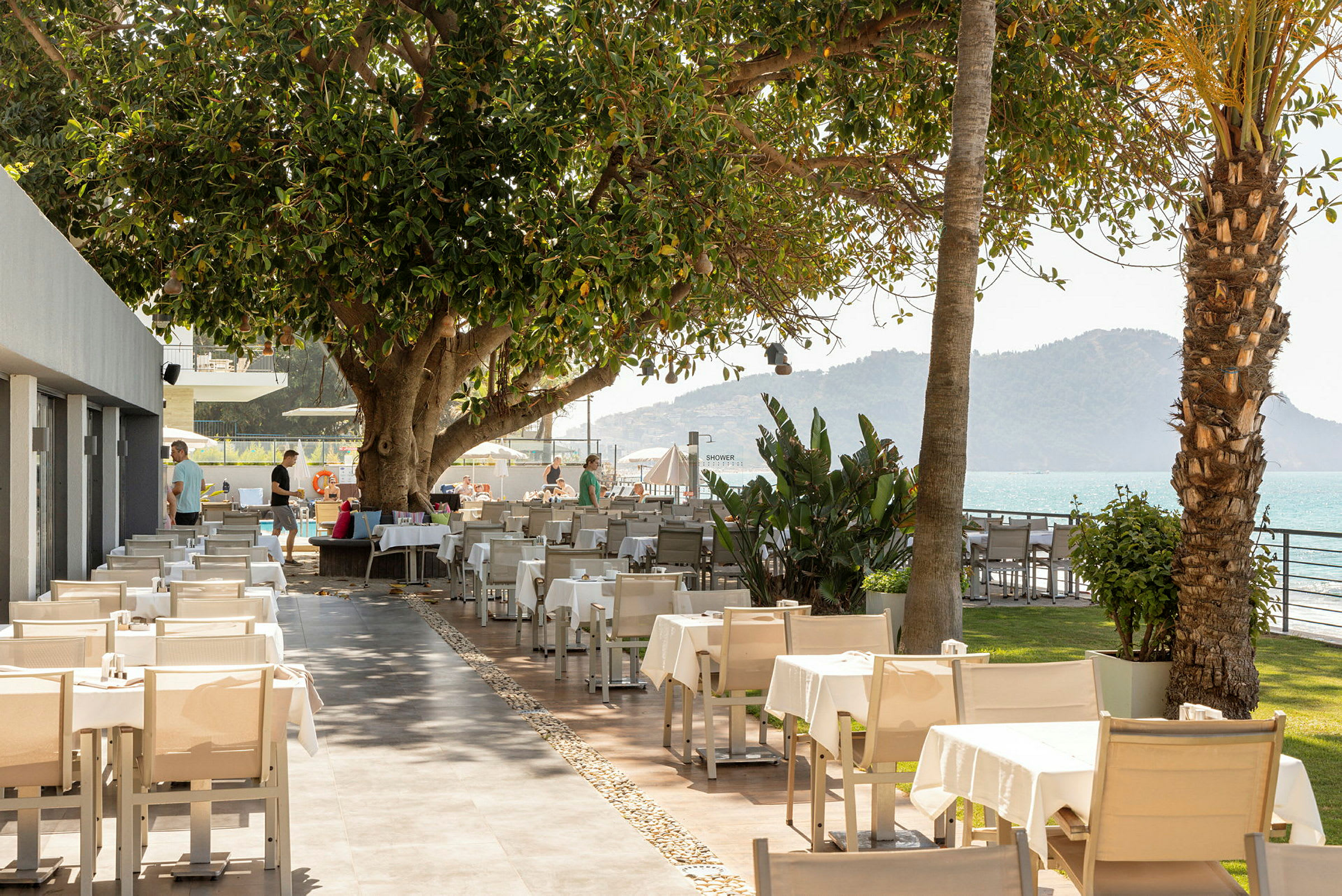 Utomhusrestaurang med sittplatser, stora träd och utsikt över havet och bergen. (AI-genererad text)