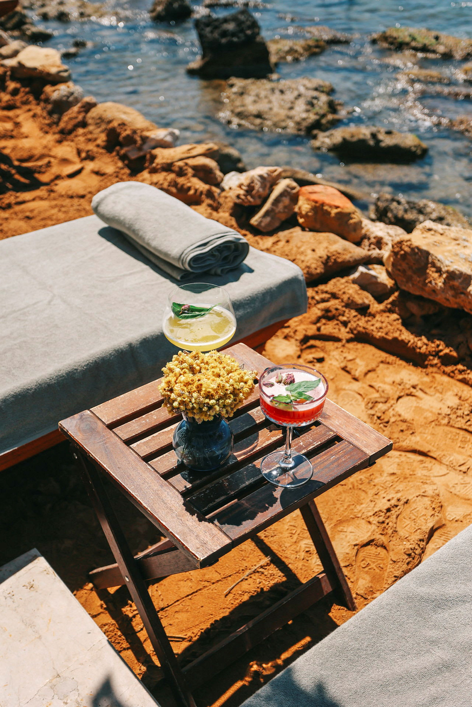 En strandscen med en hopfällbar träbord med två dekorativa drinkar och en blomvas. Bakgrunden visar havsvatten och stenig kust. (AI-genererad text)