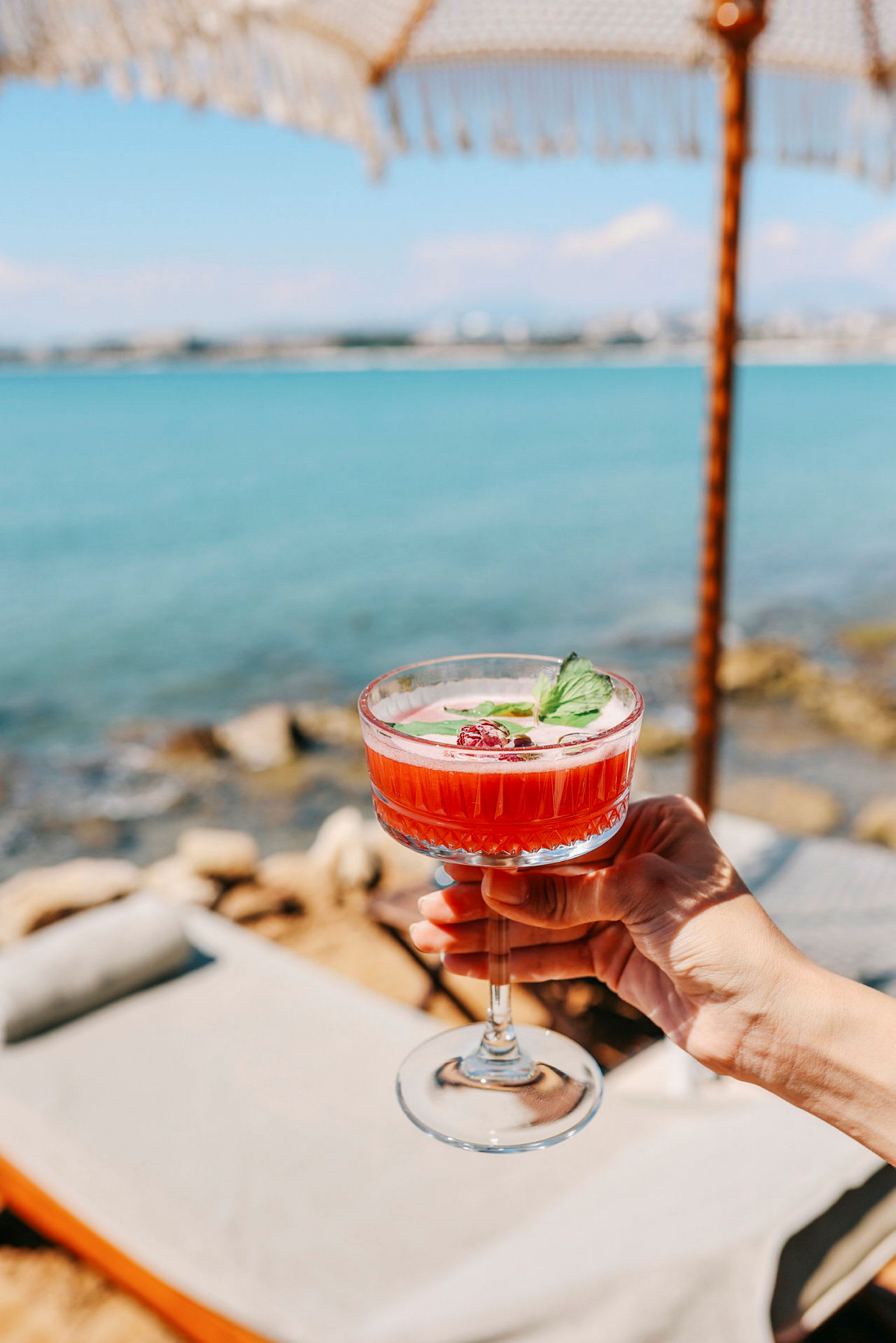 Hand håller ett glas drink med rödfärgad dryck och garnering vid strandmiljö. (AI-genererad text)