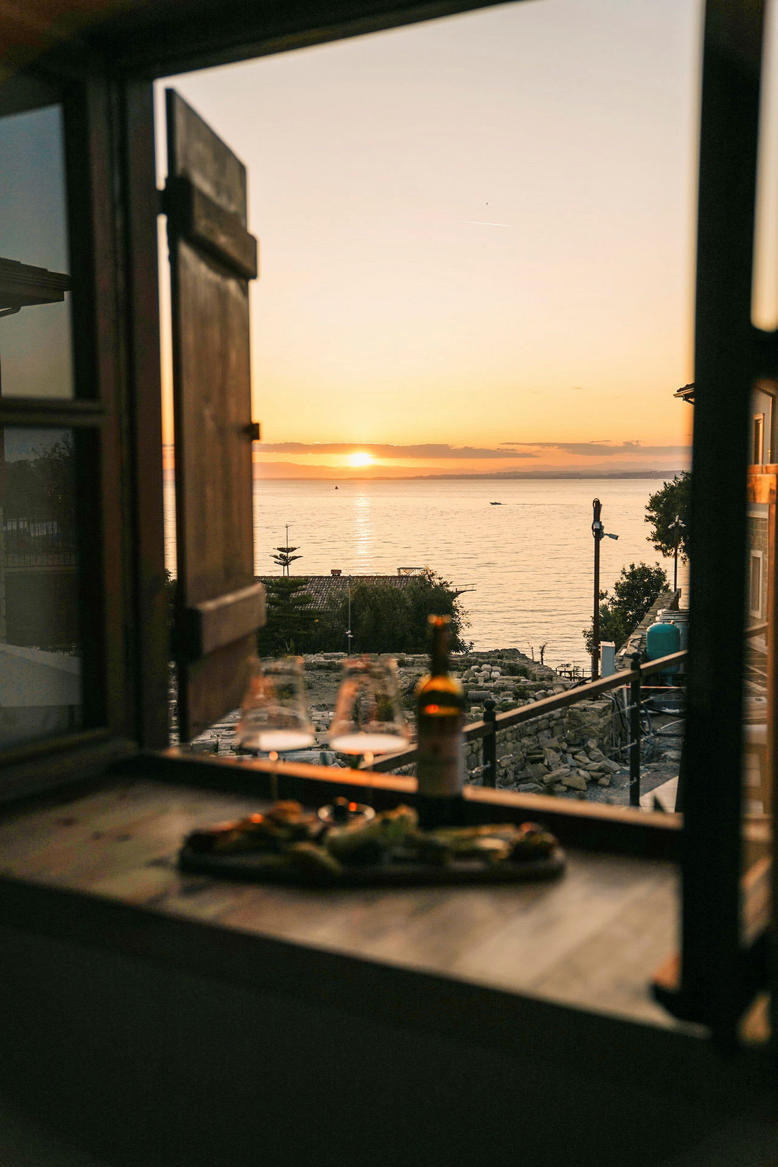 Utsikt från ett fönster mot en solnedgång över havet, med glas och en flaska på en bänk. (AI-genererad text)