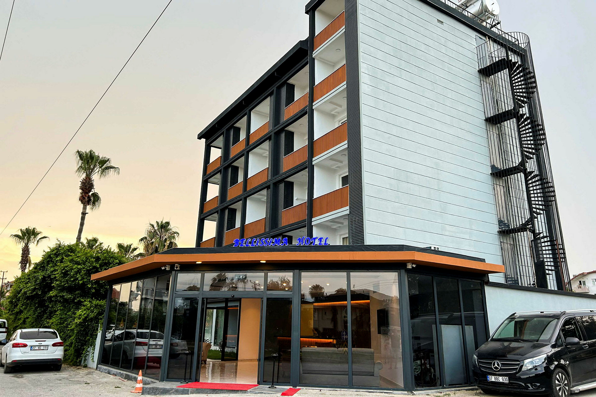 Byggnaden är ett modernt hotell med fem våningar och glasentré. Skylt för "Bellissima Hotel" visas på entrén. (AI-genererad text)