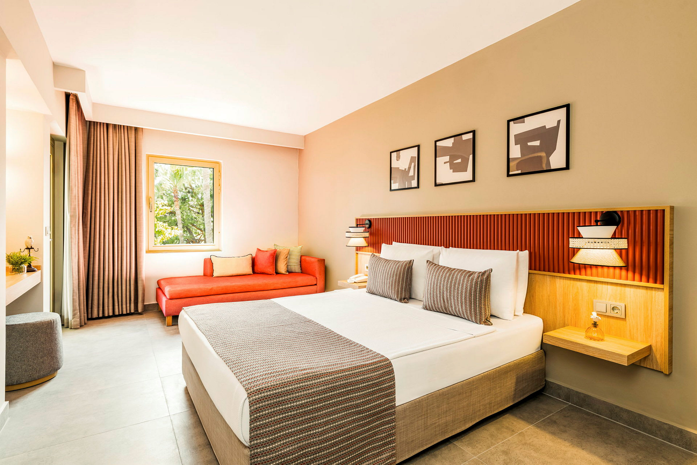 Ett modernt hotellrum med en bred säng klädd i vita lakan och dekorativa kuddar. Det finns en röd soffa vid fönstret och tre konstverk på väggen ovanför sängen. (AI-genererad text)