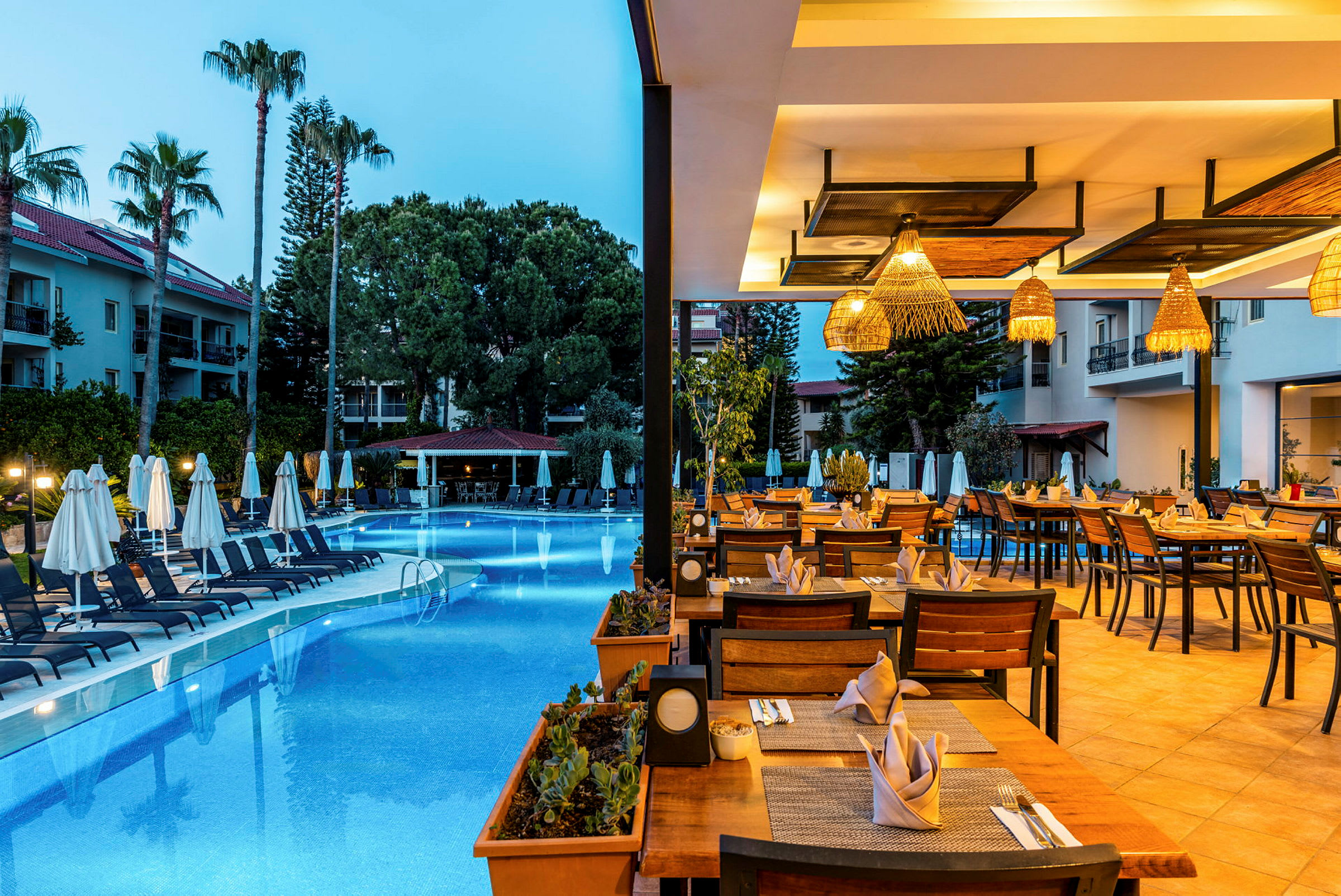 En utomhusrestaurang med bord dukade för middag bredvid en pool och omgiven av palmer. (AI-genererad text)