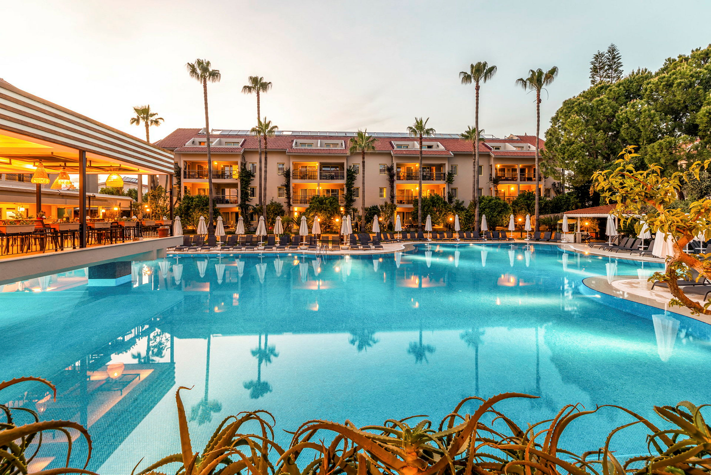 Ett hotellområde med en stor pool, omgiven av solsängar och parasoller, med byggnader i bakgrunden. (AI-genererad text)