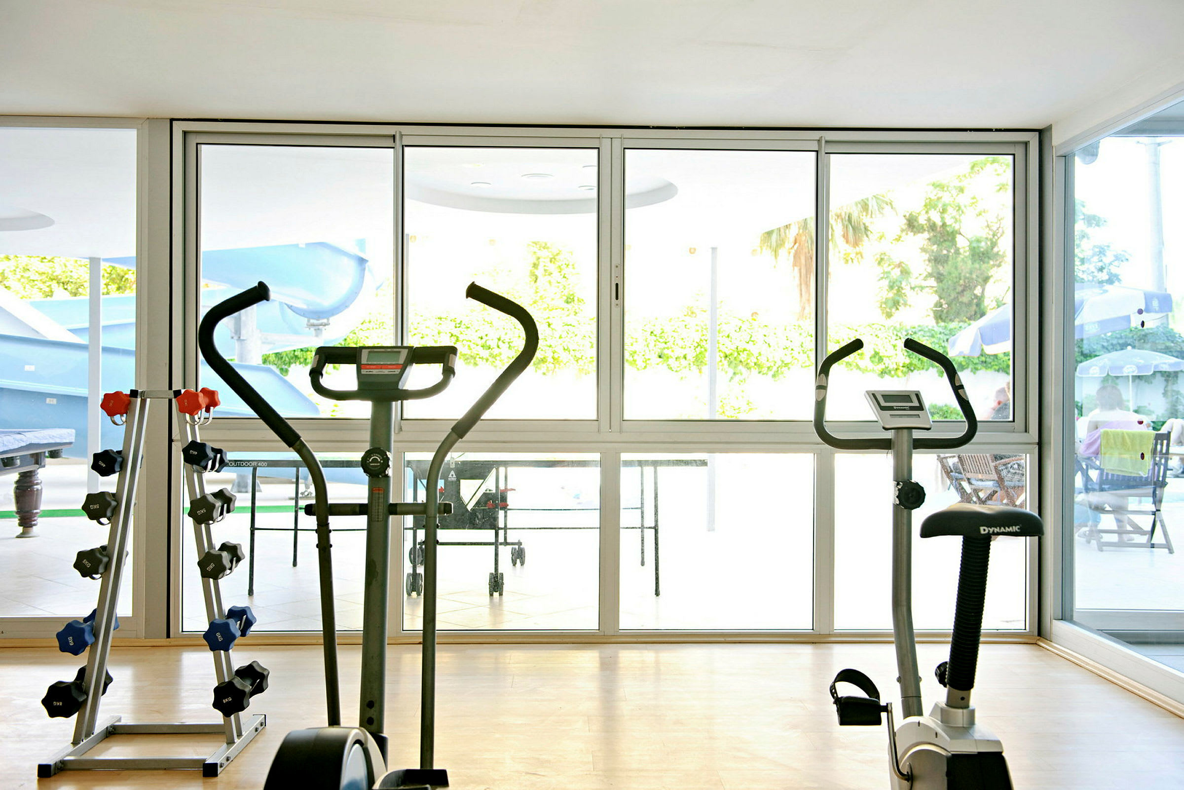 Ett gym med träningscyklar och hantlar, utsikt mot ett poolområde genom glasväggar. (AI-genererad text)