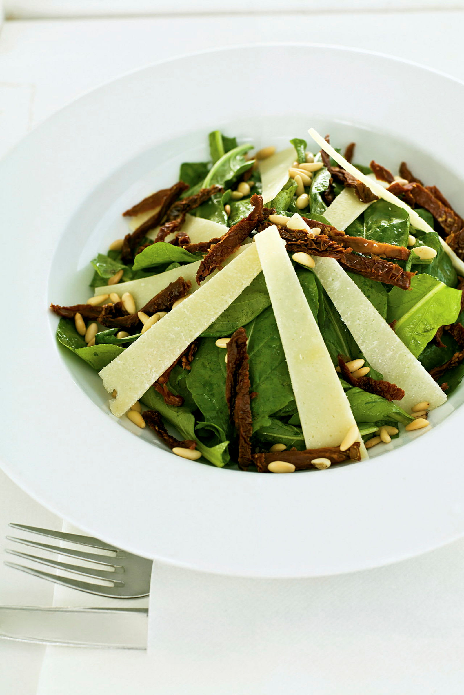 En tallrik med sallad innehållande gröna blad, soltorkade tomater, pinjenötter och tunnskivad ost. (AI-genererad text)