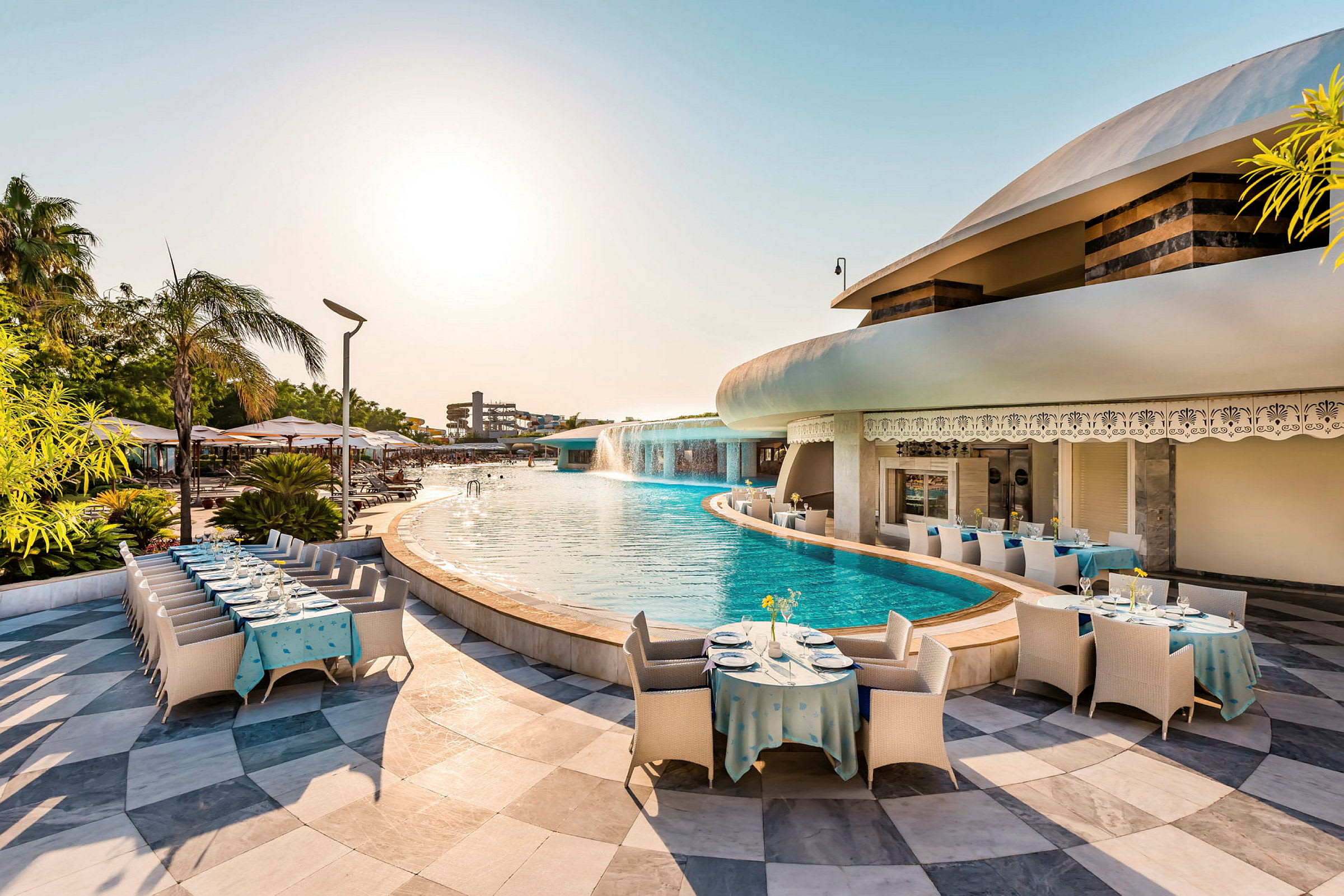Bild på en pool med vattenfall nära en restaurang med dukade bord och parasoller. (AI-genererad text)