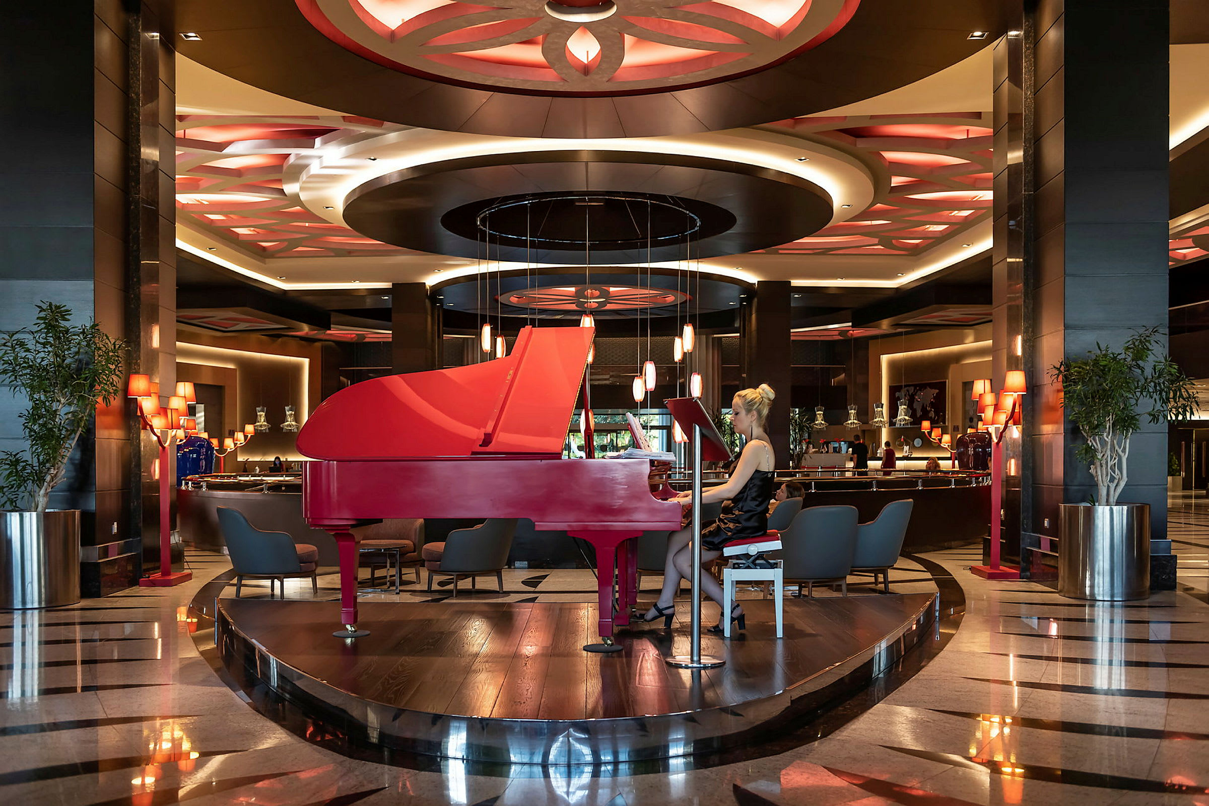 En röd flygel står centralt placerad i en elegant hotellobby med dekorativa taklampor. (AI-genererad text)