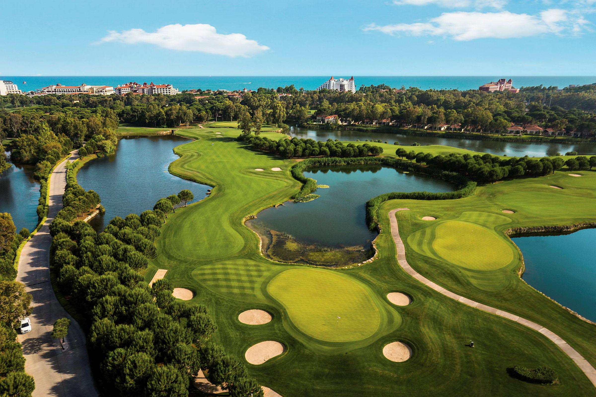 Flygfoto över en golfbana med gröna fairways, bunkrar och vattenhinder, omgiven av hotell och träd. (AI-genererad text)