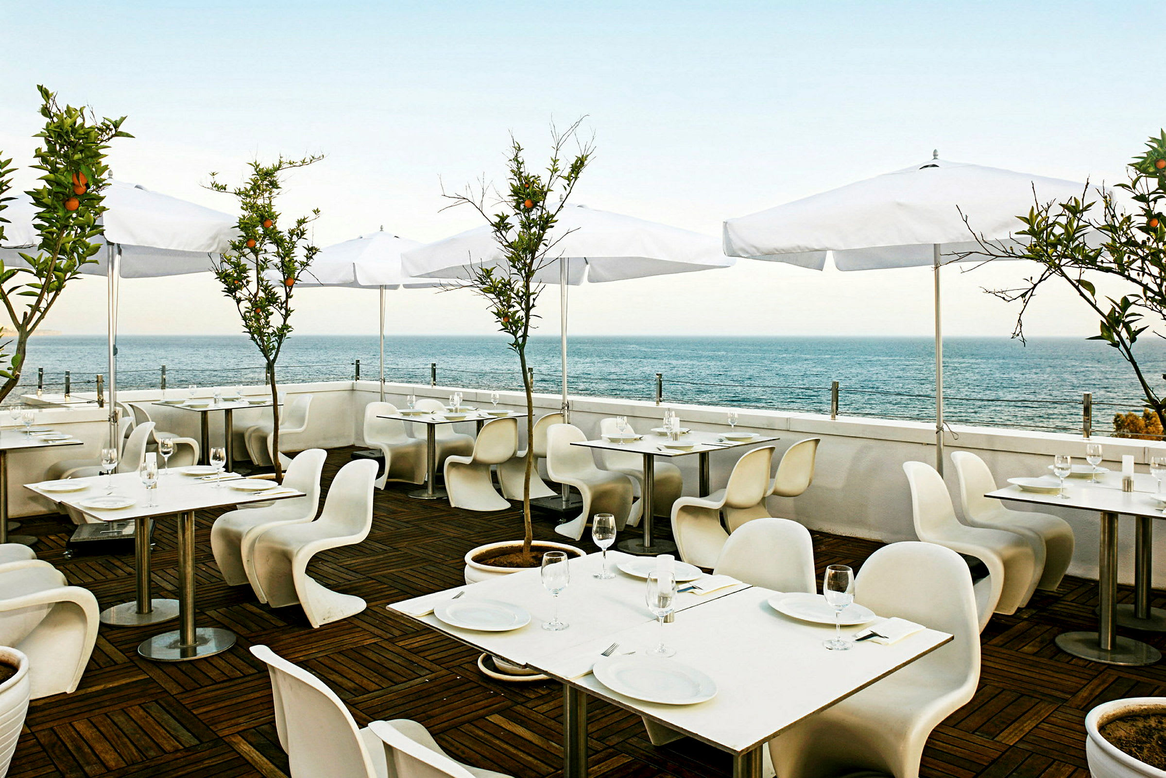 Restaurang med uteservering, vita bord och stolar samt vita parasoller. Bakgrund med utsikt över havet. (AI-genererad text)