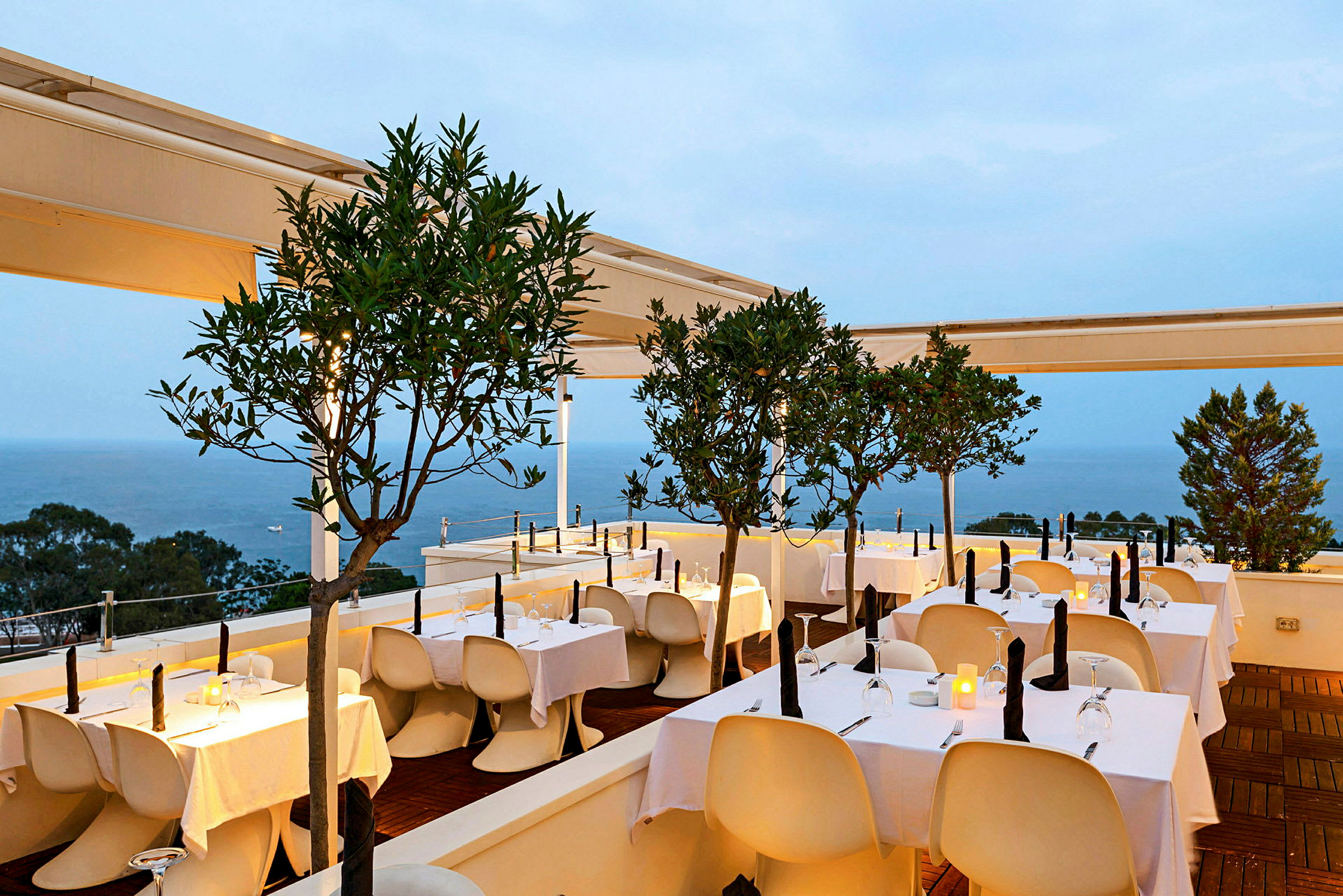 En elegant utomhusrestaurang belägen vid havet, med vita dukar på borden och små träd som dekoration. (AI-genererad text)