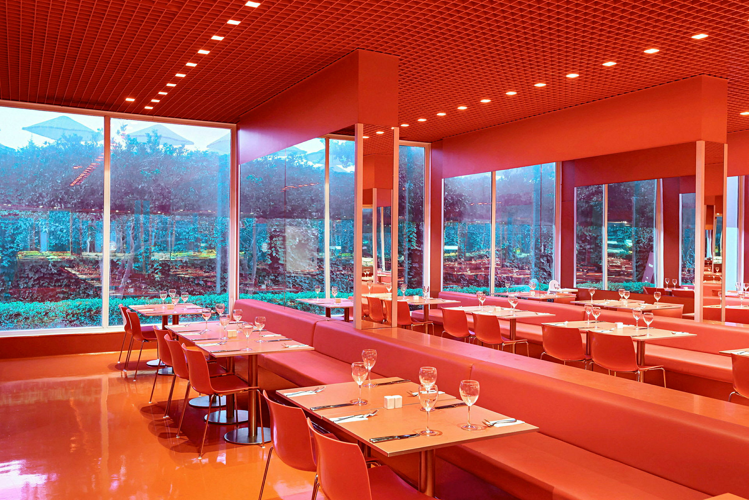 En modern restaurang med röda möbler och stora glasfönster som vetter mot en grönskande trädgård. (AI-genererad text)