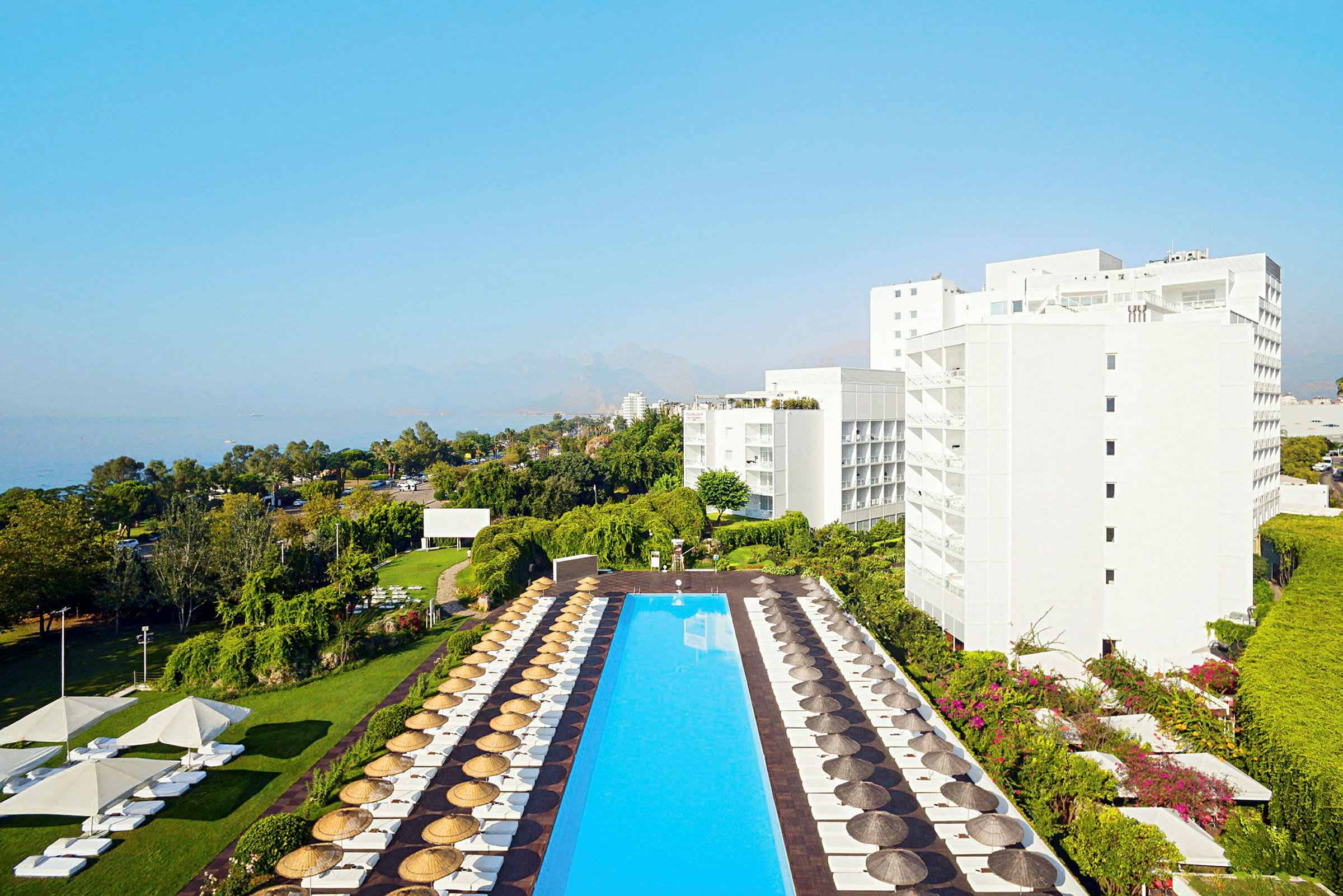 Ett hotellkomplex sett från ovan med en stor rektangulär pool omgiven av solstolar och parasoller. Träd och grönska omger området, och i bakgrunden syns havet och berg. (AI-genererad text)