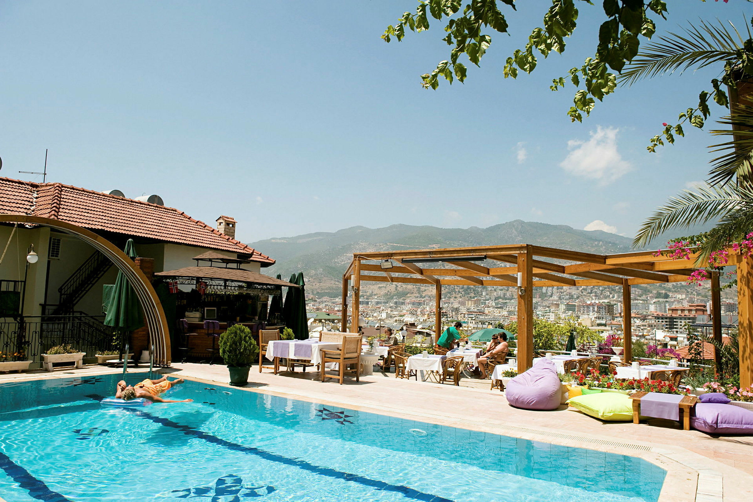 En utomhuspool vid ett hotell med solstolar och parasoller. En träpergola och utsikt över en stad och berg i bakgrunden. (AI-genererad text)