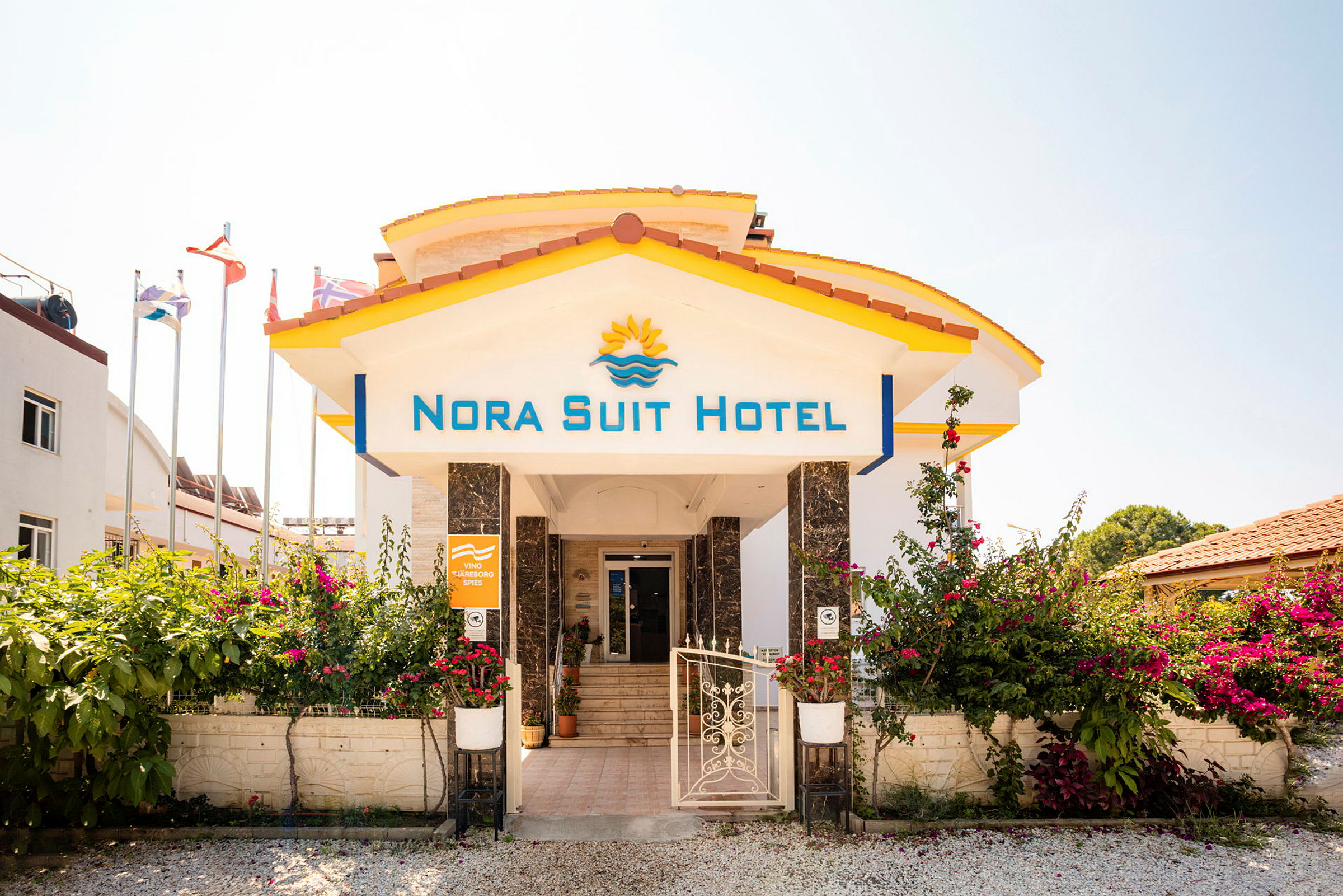 Entrén till Nora Suit Hotel med blommor och flaggor i bakgrunden. (AI-genererad text)