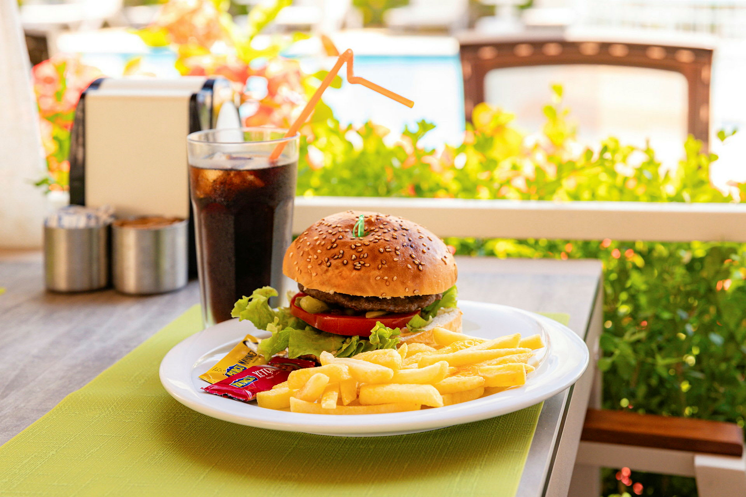 En tallrik med en hamburgare, pommes frites och kryddpåsar, samt ett glas läsk, står på ett bord utomhus. (AI-genererad text)