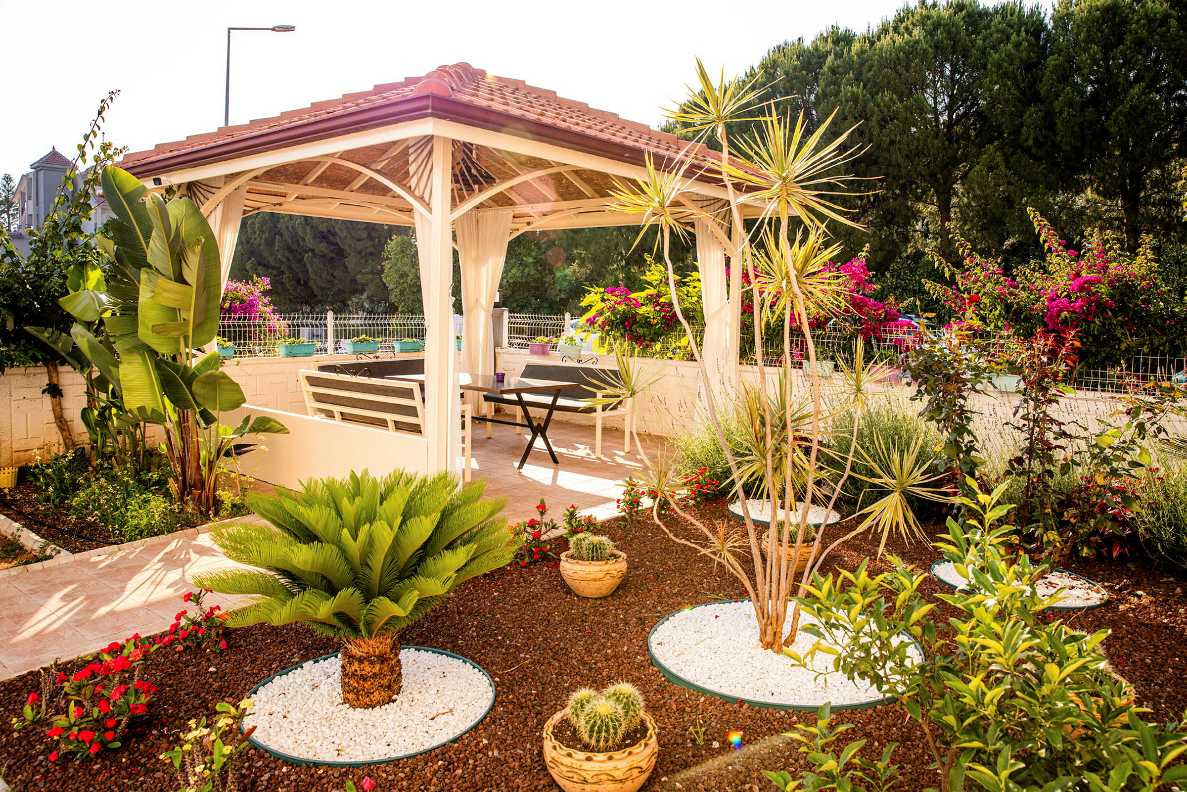 En trädgård med en pergola omgiven av exotiska växter och blommor. Under pergolan finns möbler för avkoppling. (AI-genererad text)