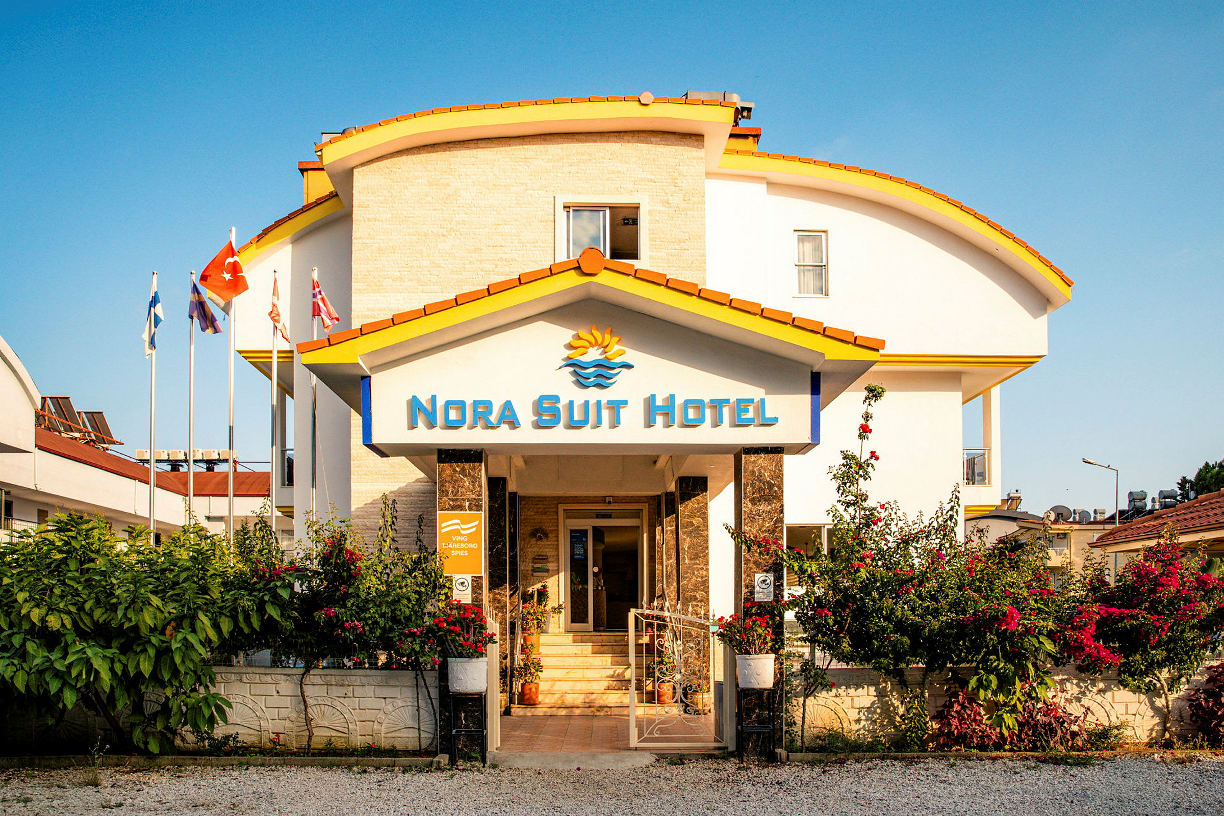 Byggnaden till Nora Suit Hotel med en ingång omgiven av blommor och flaggor. (AI-genererad text)