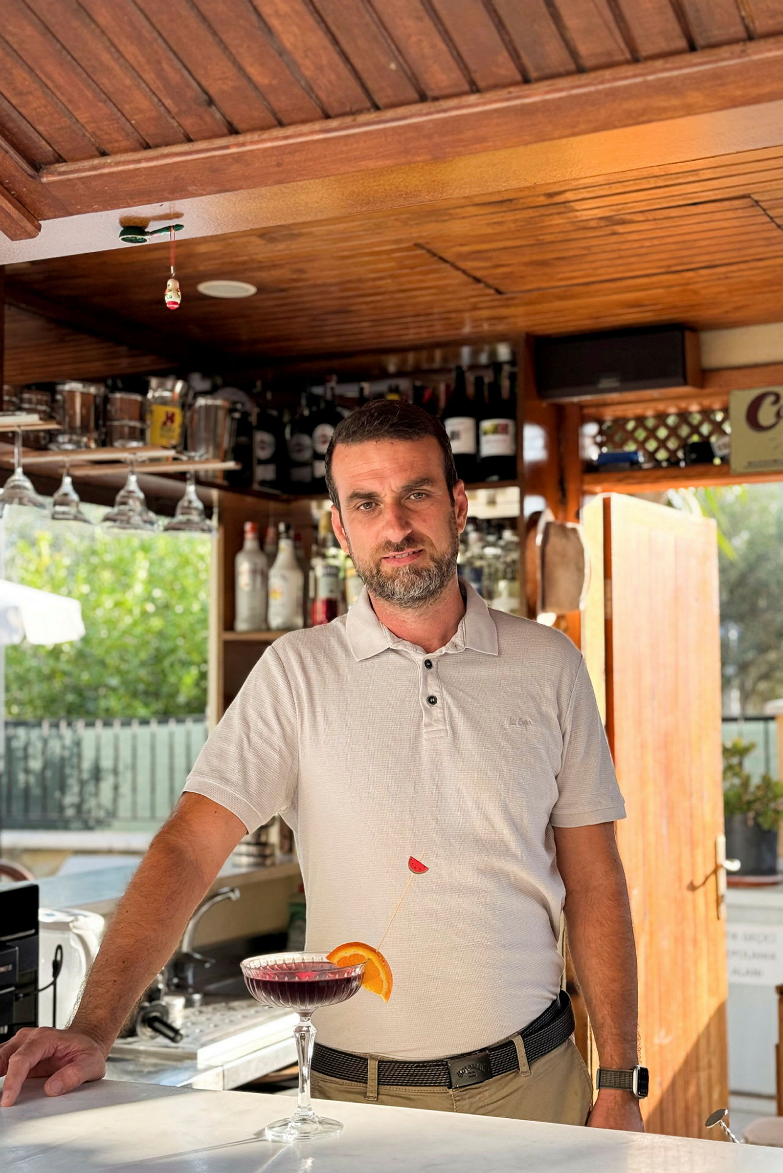 En vit skjorta vid en bar med hyllor fyllda av flaskor i bakgrunden, samt en cocktailglas med en färgad dryck framför sig. (AI-genererad text)