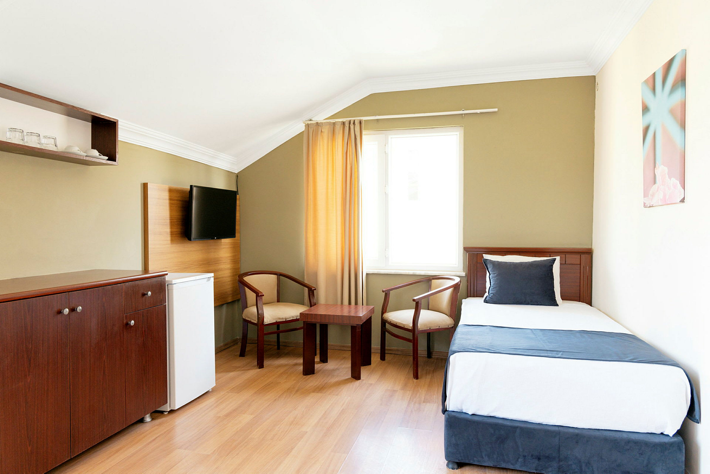 Ett hotellrum med en enkelsäng, två stolar och ett mindre bord, samt en väggmonterad TV. (AI-genererad text)