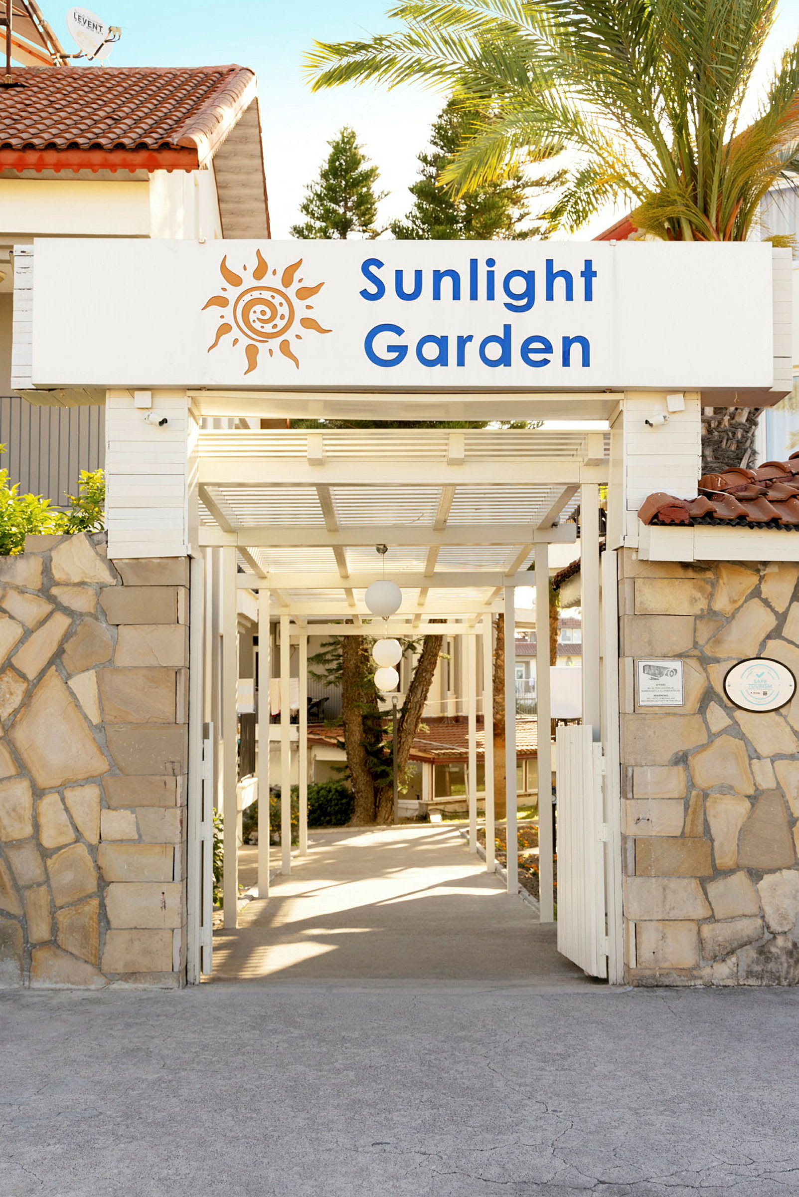 Entré med skylt som lyder 'Sunlight Garden', omgiven av stenmur och tropiska träd. (AI-genererad text)