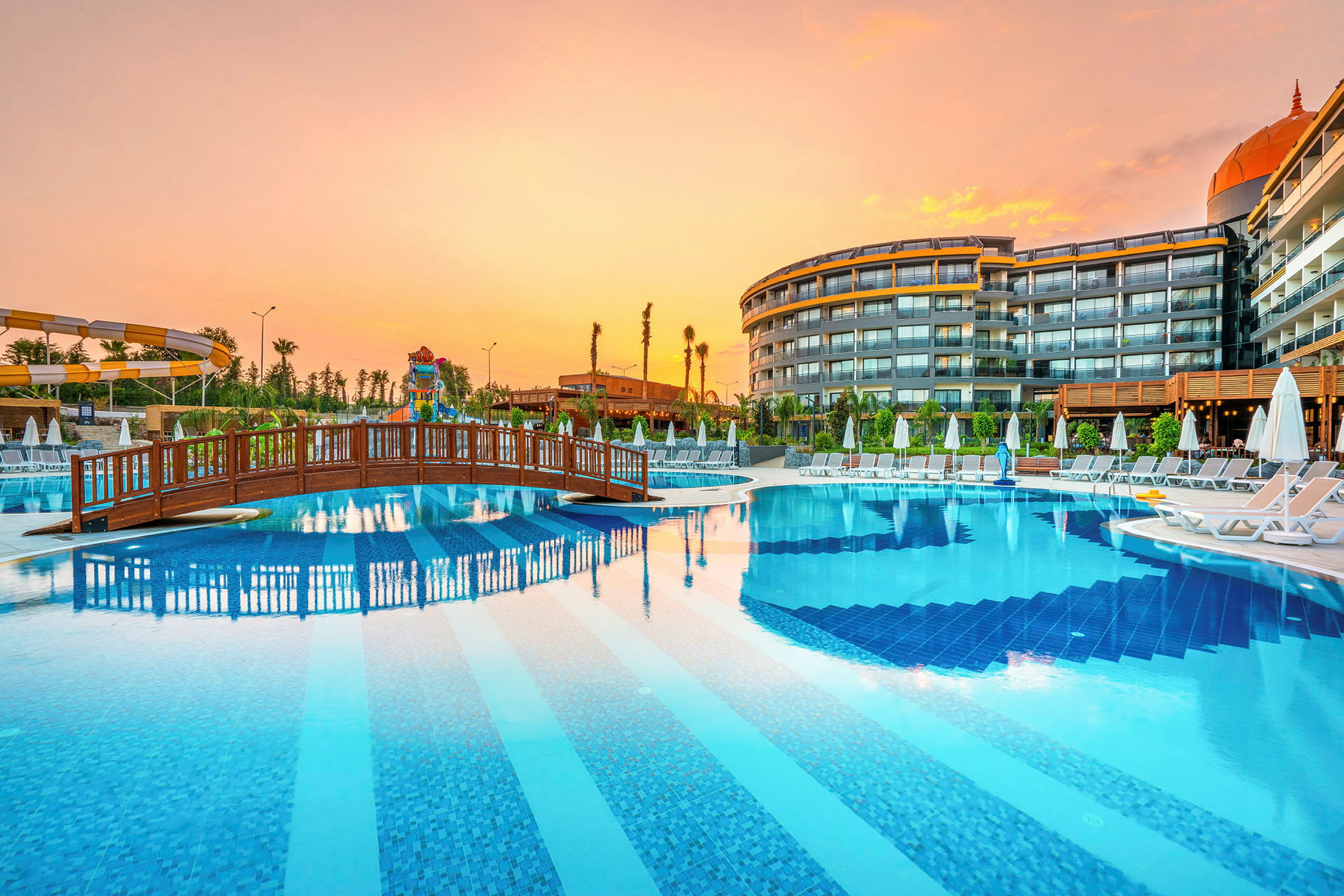 En hotellpool med bro och solstolar, omgiven av byggnader vid solnedgång. (AI-genererad text)