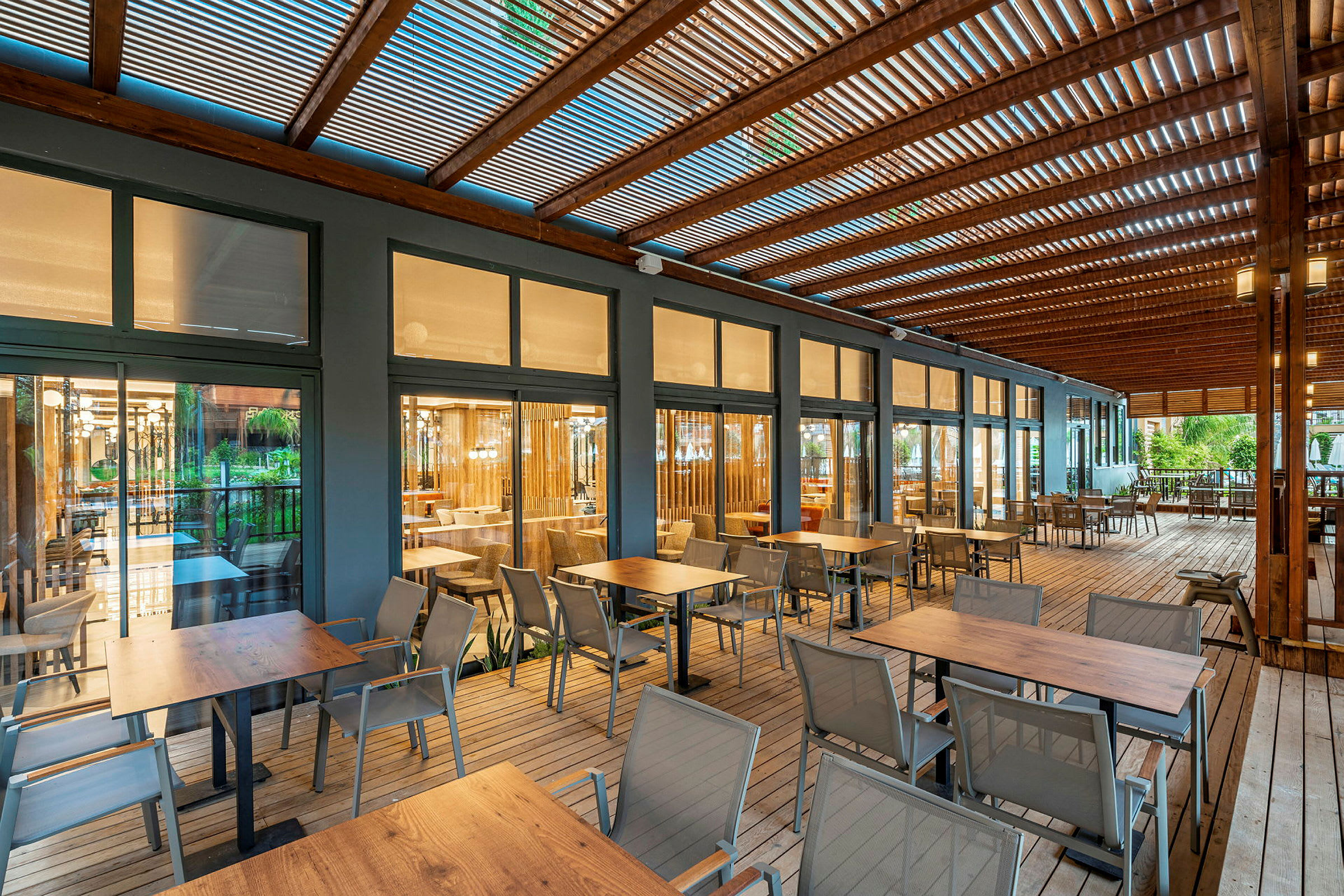 En uteplats med bord och stolar, omgiven av trästruktur och glasväggar som vetter mot en inomhusrestaurang. (AI-genererad text)
