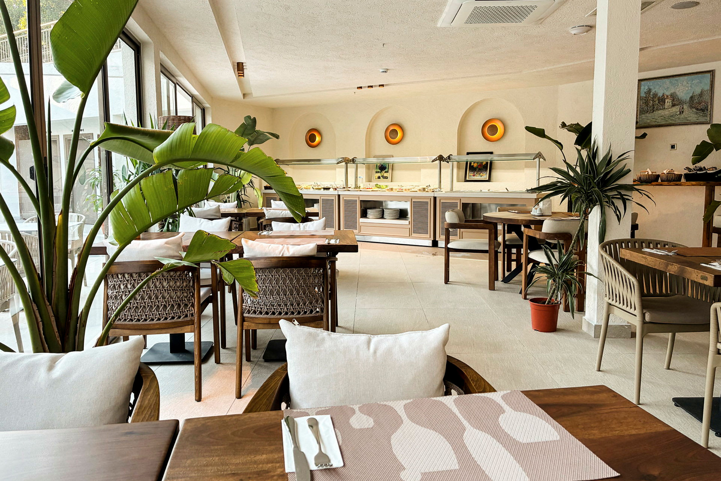 En ljus och inbjudande restaurang med trämöbler, grön växtinredning och ett bufféområde i bakgrunden. (AI-genererad text)
