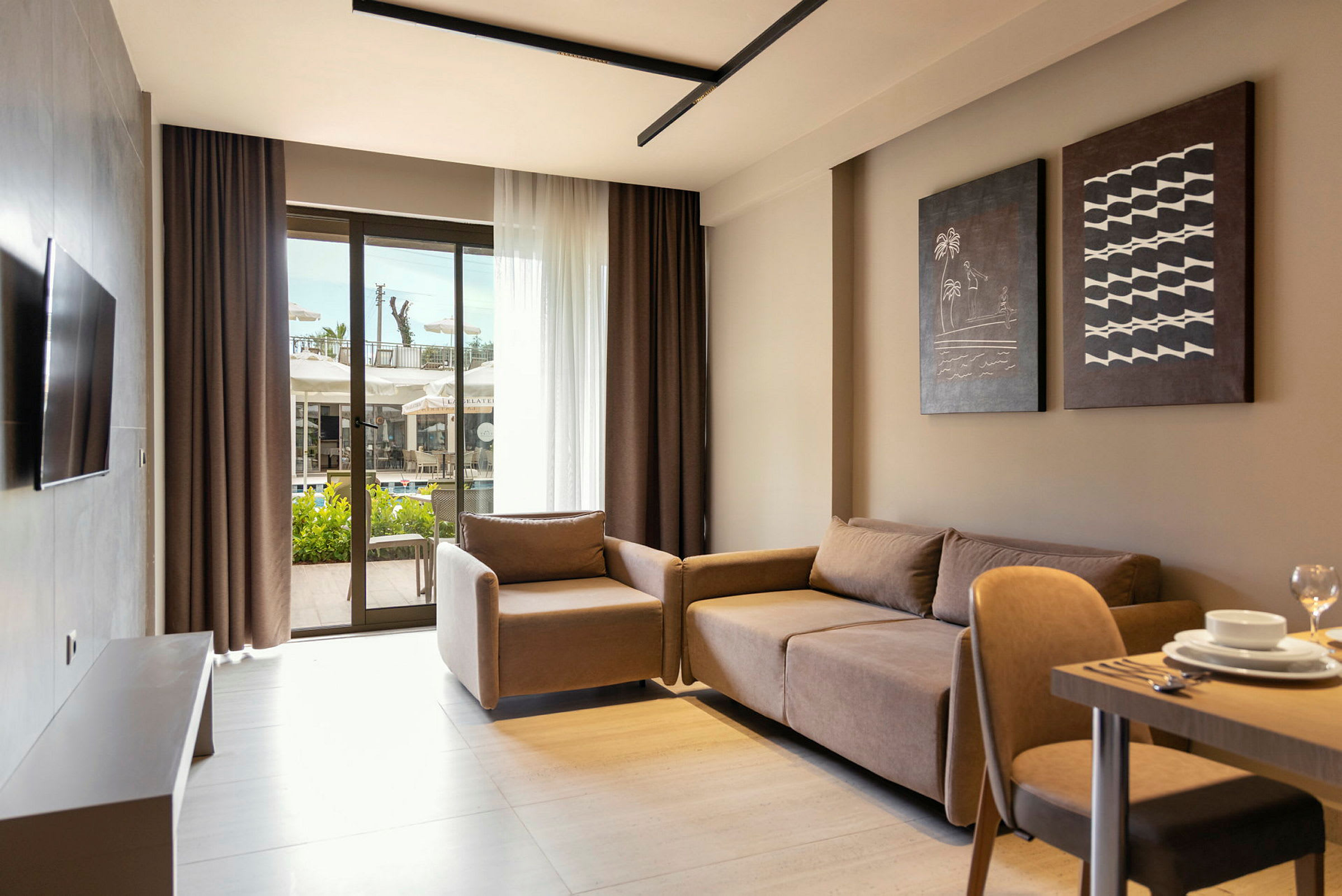 Ett modernt hotellrum med soffa, fåtölj och bord sett från inomhus. Stora glasdörrar i bakgrunden visar utsikten mot en terrass och swimmingpool. (AI-genererad text)