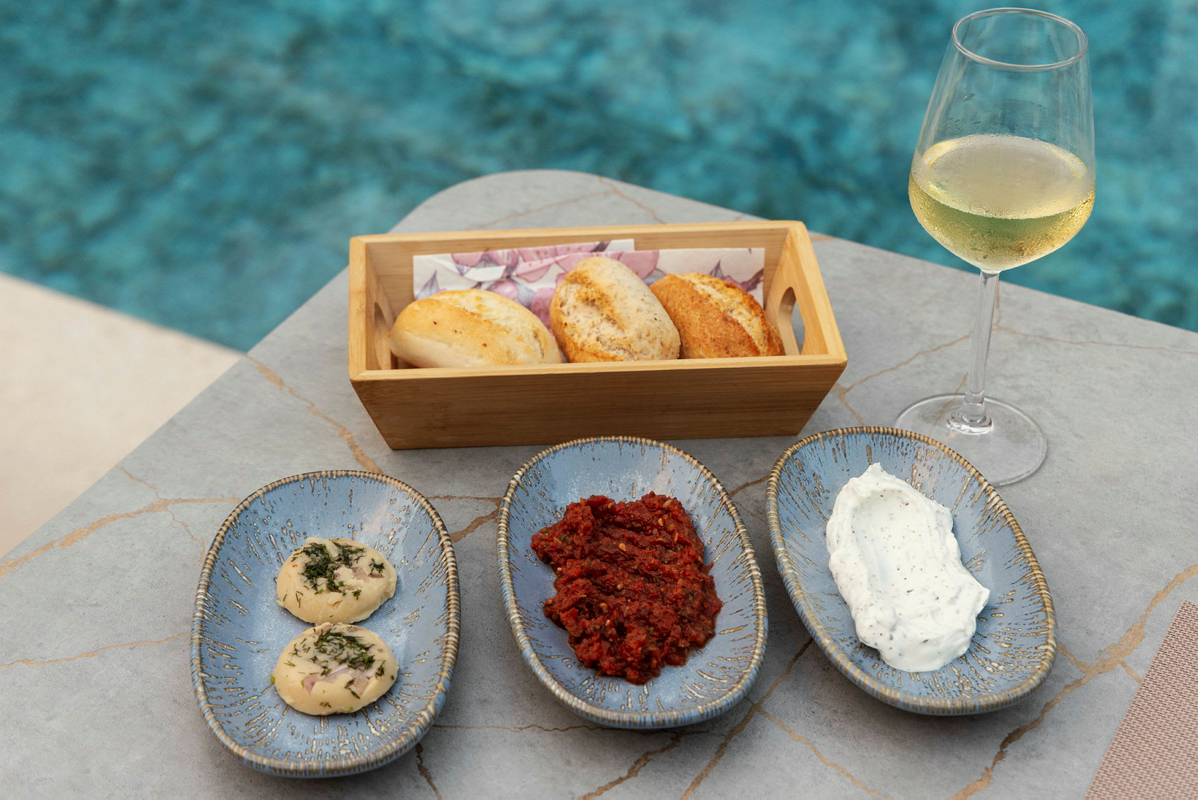 En träkorg med bröd, tre keramikfat med tillbehör och ett glas vitt vin stående vid en pool. (AI-genererad text)
