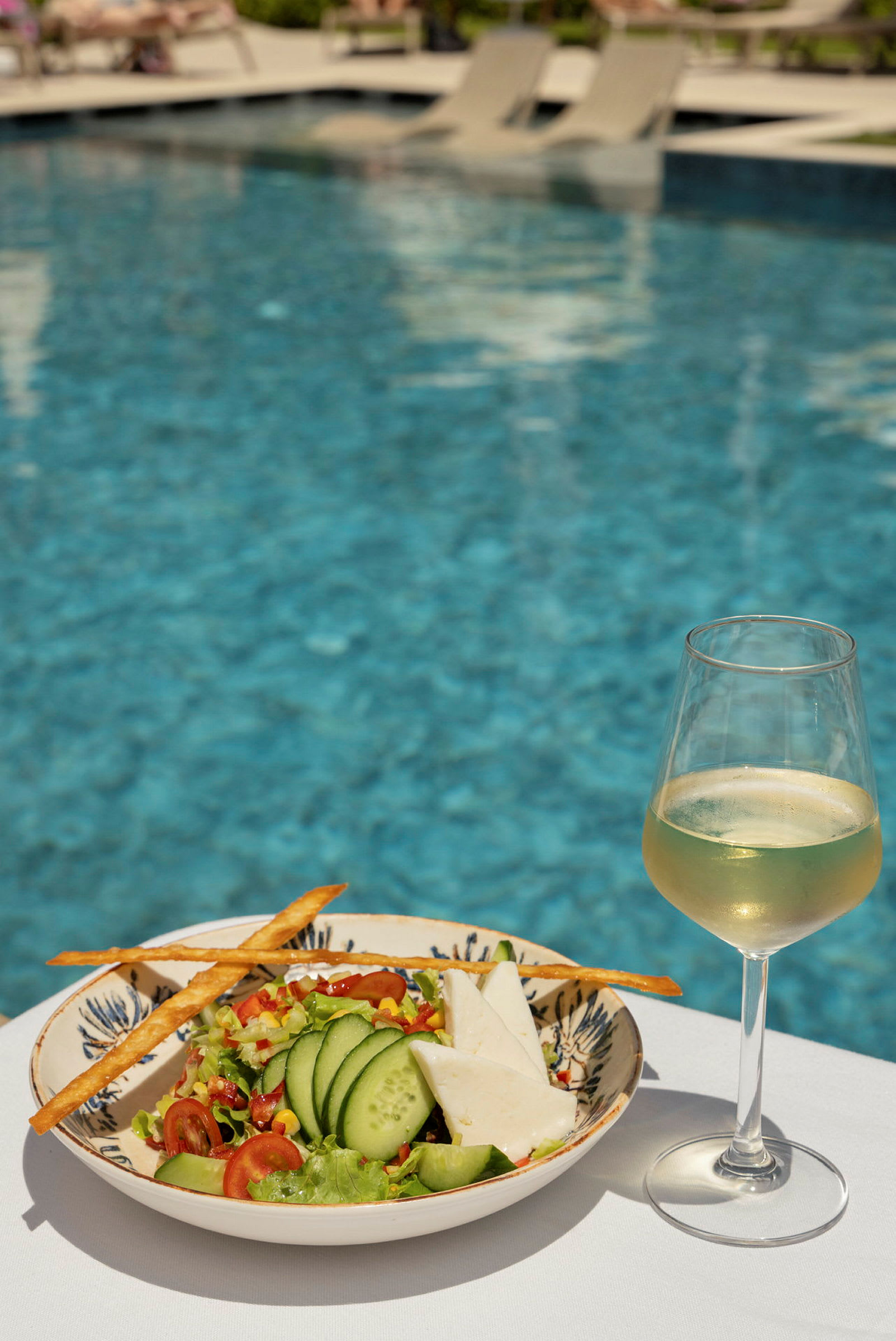 En sallad med gurka och ost samt ett glas vitt vin står på ett bord med en pool i bakgrunden. (AI-genererad text)