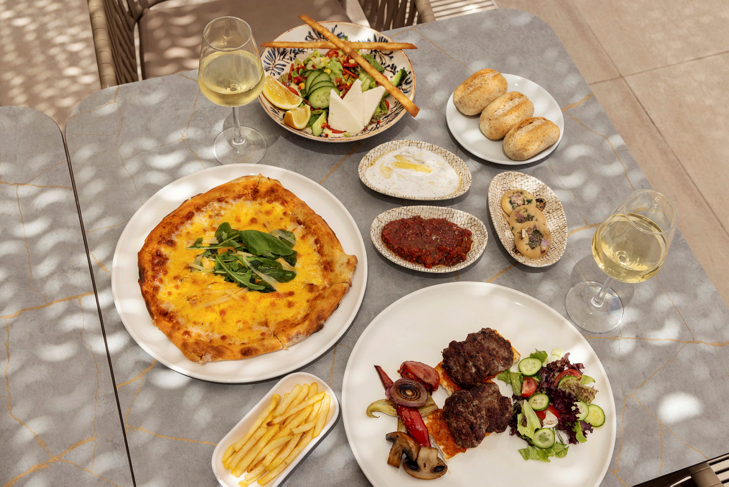 Ett bord med olika maträtter som inkluderar pizza, sallad, bröd, kött med grönsaker, pommes frites samt glas med vitt vin. (AI-genererad text)