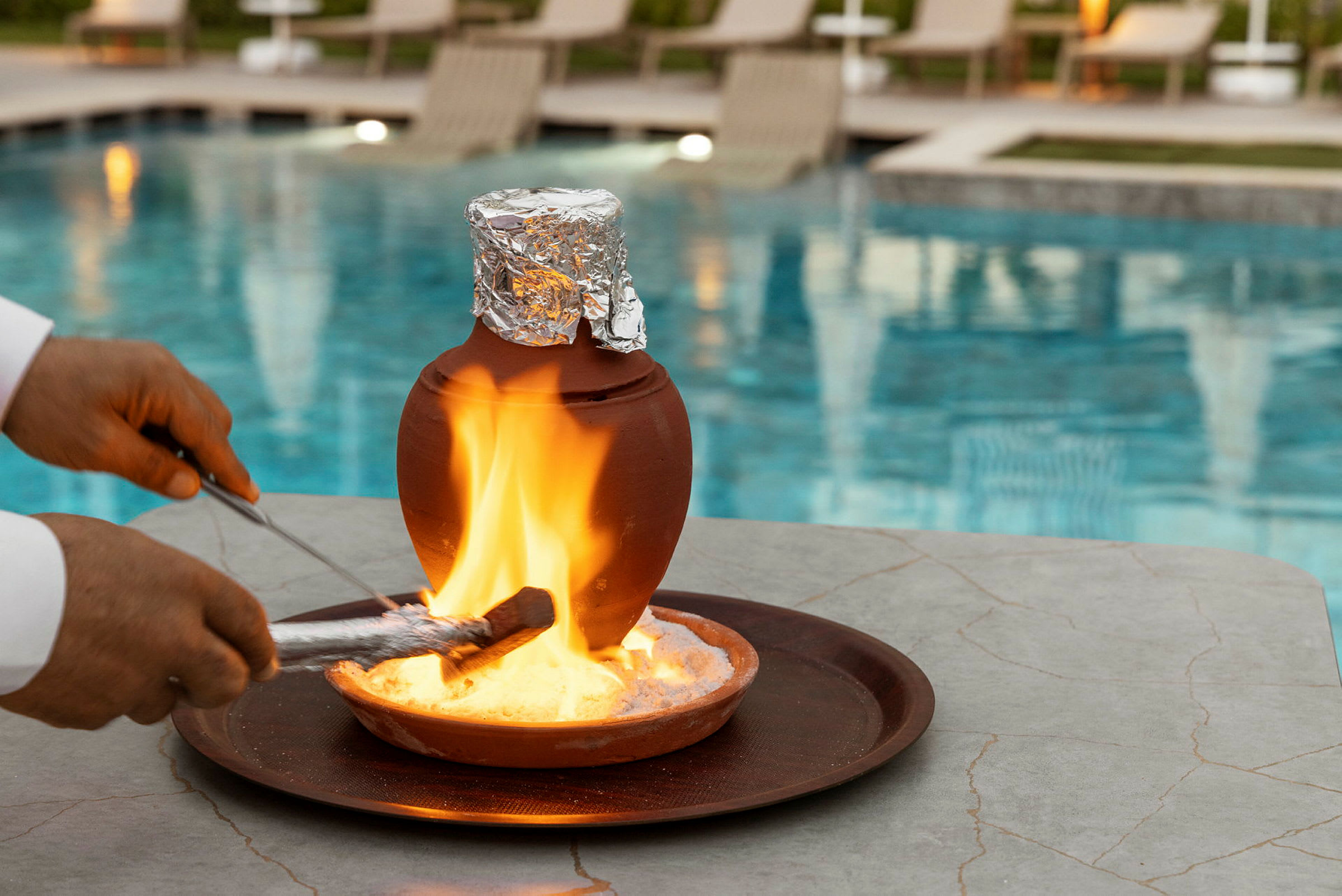 Ett bord med en lerkruka täckt av folie står ovanpå en bricka, med en eld tänd nedanför. I bakgrunden syns en pool och solstolar. (AI-genererad text)