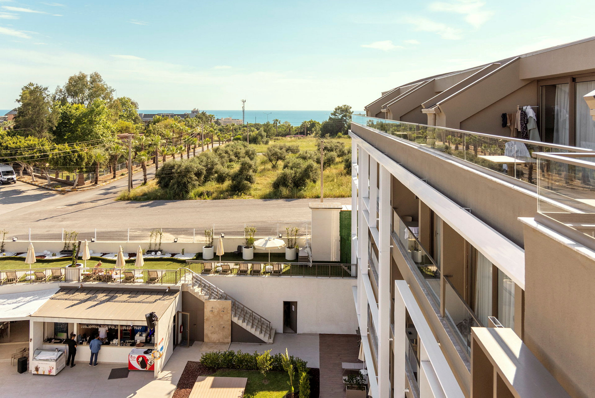 Ett hotell med modern arkitektur och balkonger, bredvid ett trädbevuxet område och en väg. I bakgrunden kan havet ses. (AI-genererad text)