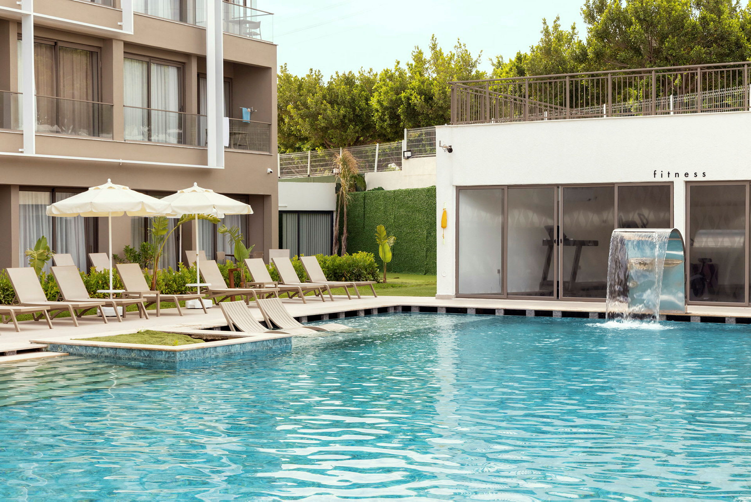 Utomhuspool med solstolar och parasoller vid ett hotell eller resort. En vattenfallfunktion finns vid poolen och ett fitnessrum syns i bakgrunden. (AI-genererad text)