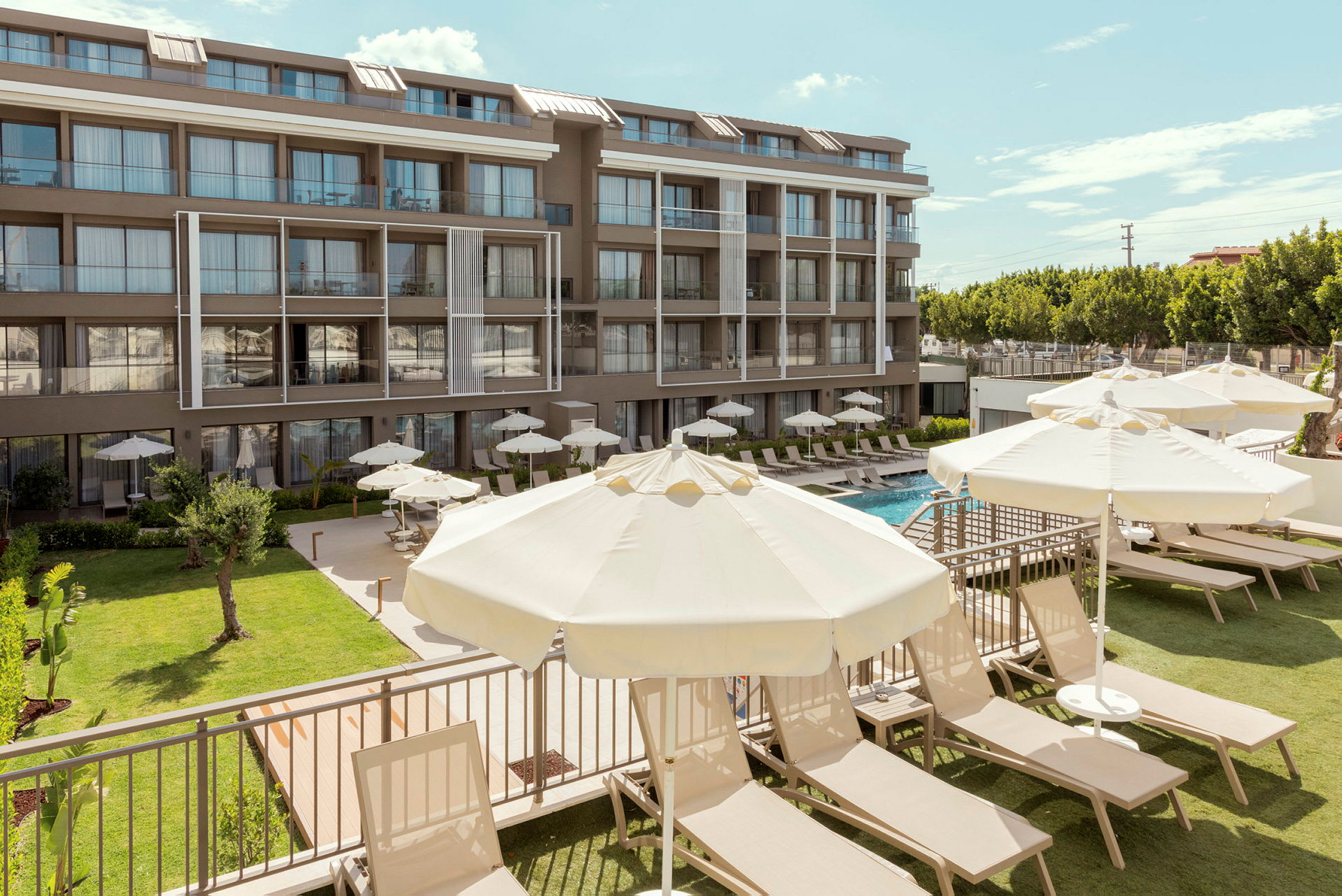 En hotellbyggnad med flera våningar i modern stil. Framför byggnaden finns en gräsmatta, solsängar och parasoller vid en swimmingpool. (AI-genererad text)