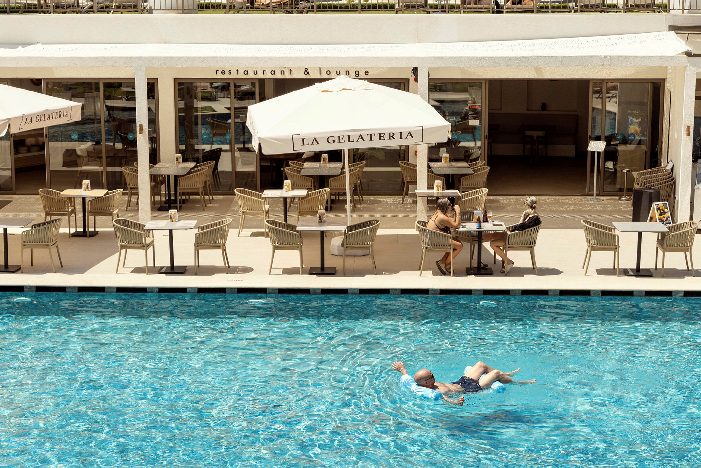 En person flyter i en pool framför en restaurangterrass med parasoller och sittplatser. (AI-genererad text)