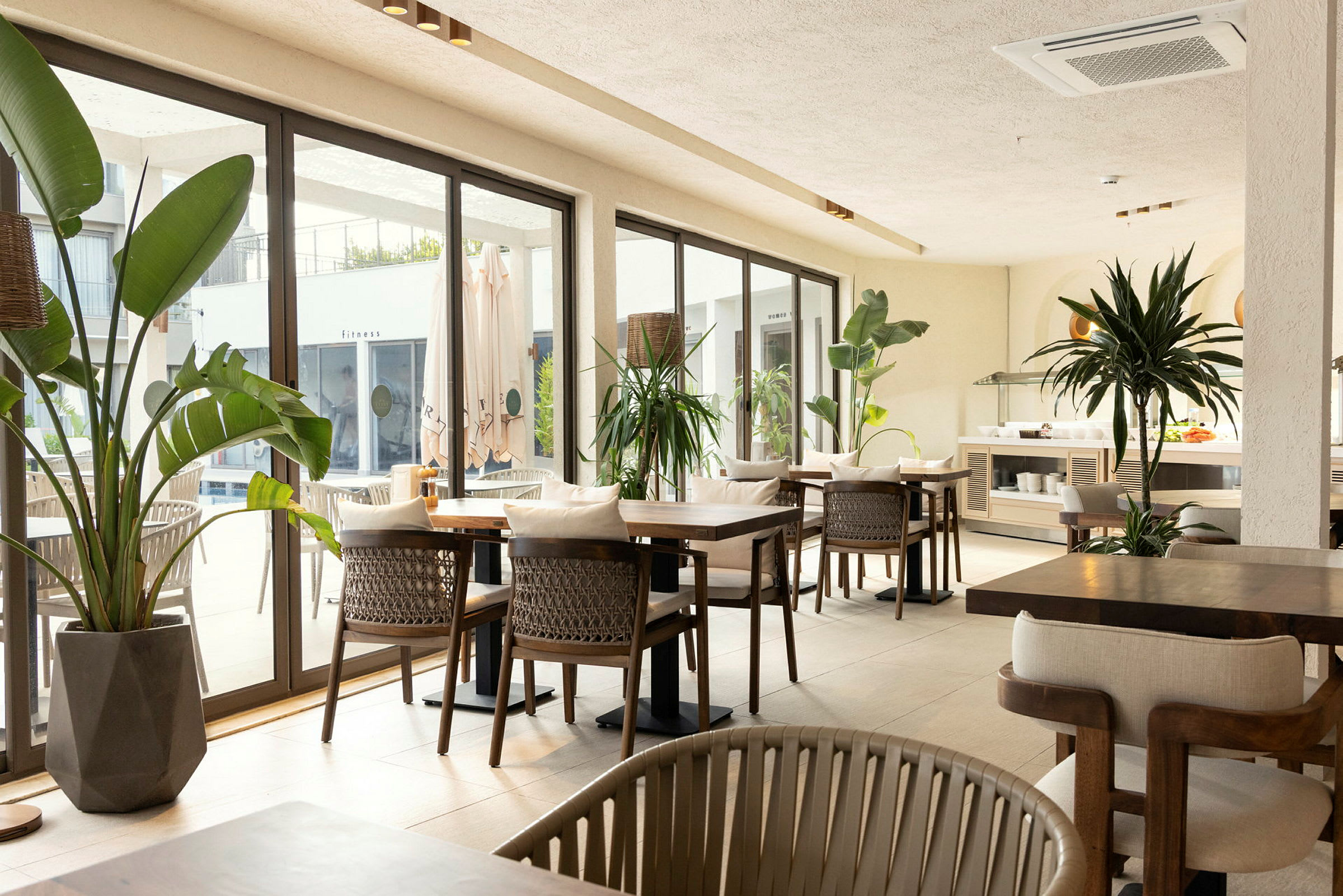 Restaurangområde med bord och stolar, omgiven av stora fönster och gröna växter. (AI-genererad text)