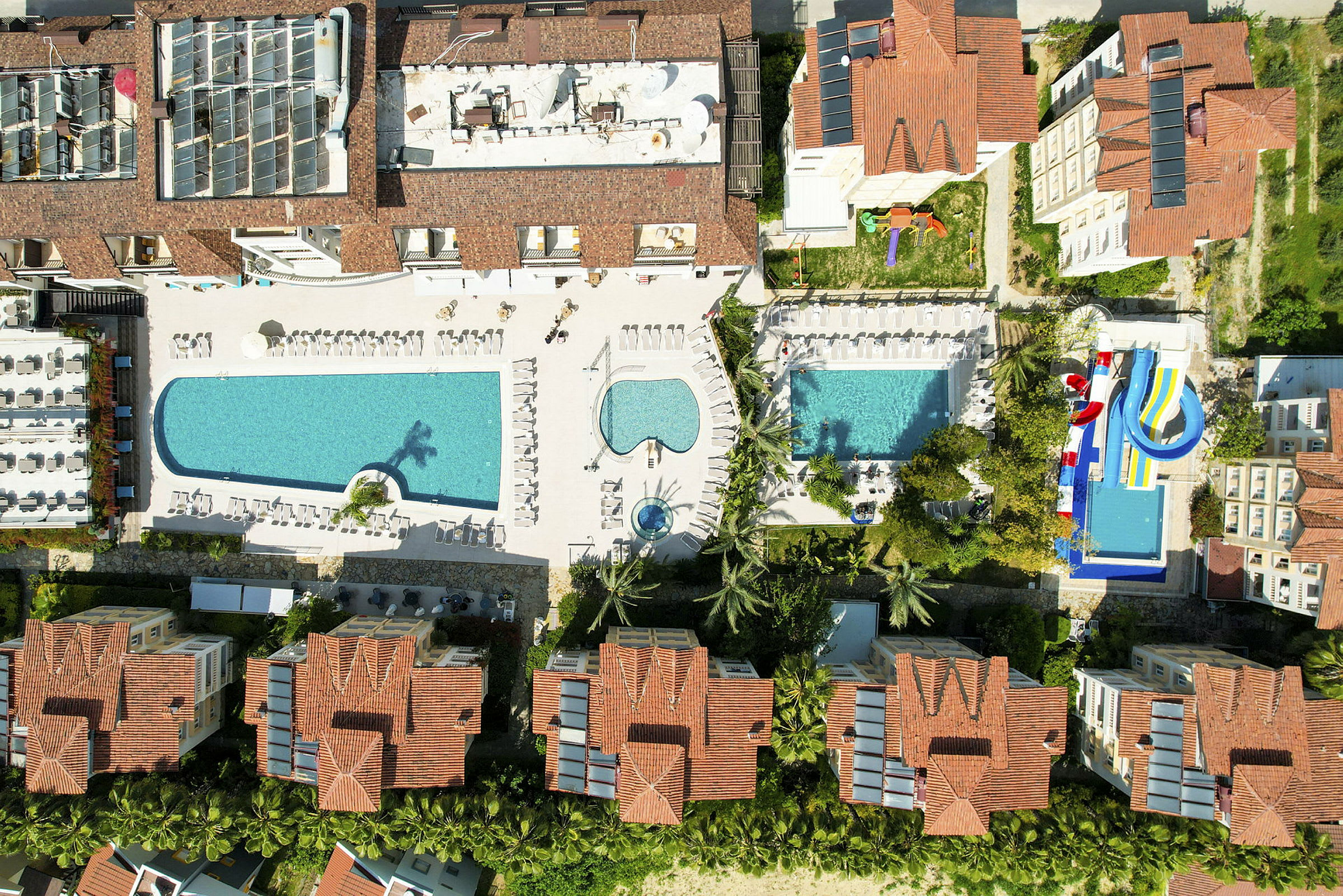 Flygfoto av hotellkomplex med pooler och vattenrutschbanor, omgivna av byggnader med röda tak. (AI-genererad text)