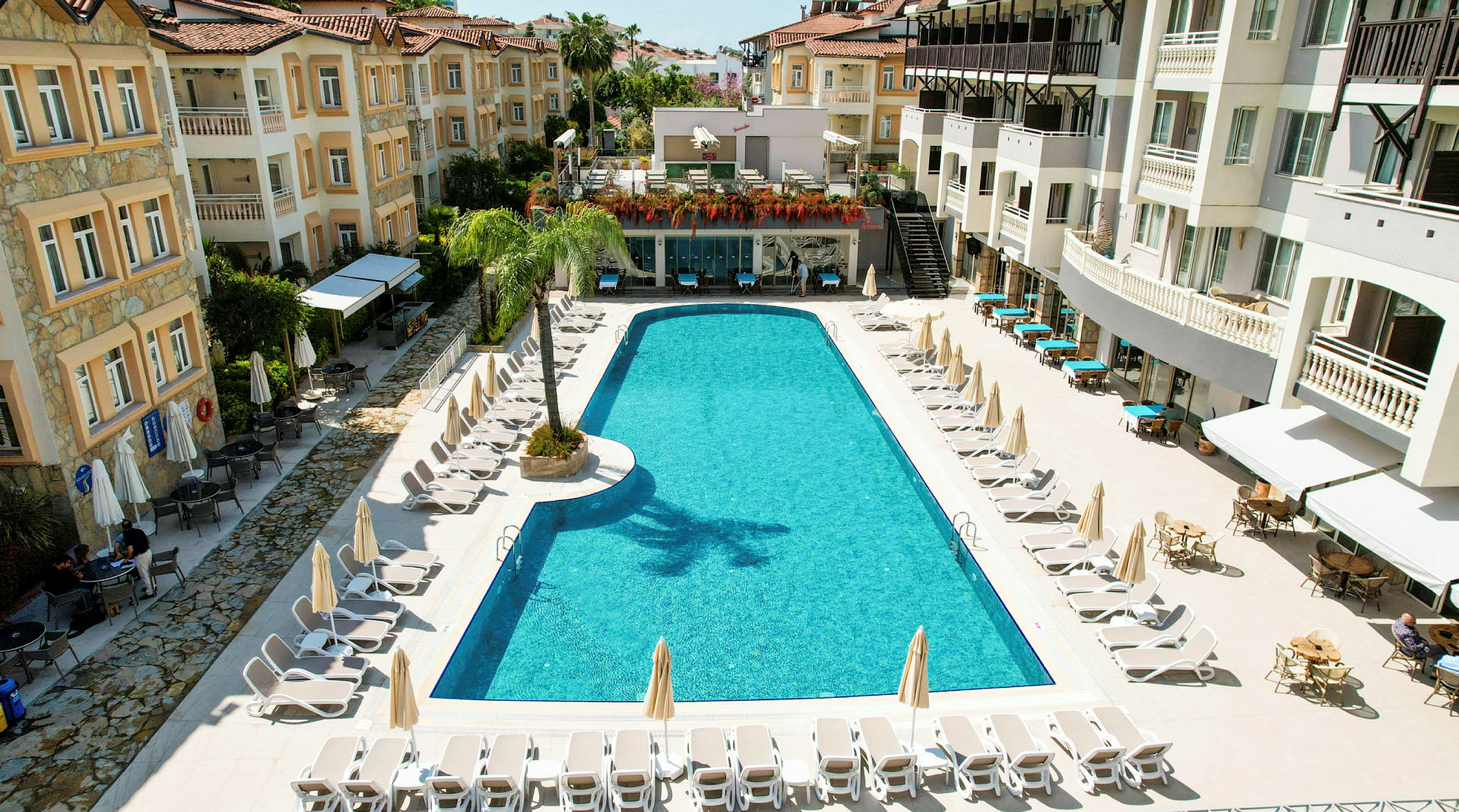 Hotellområde med lång swimmingpool omgiven av solstolar och parasoller, omgiven av byggnader med balkonger. (AI-genererad text)