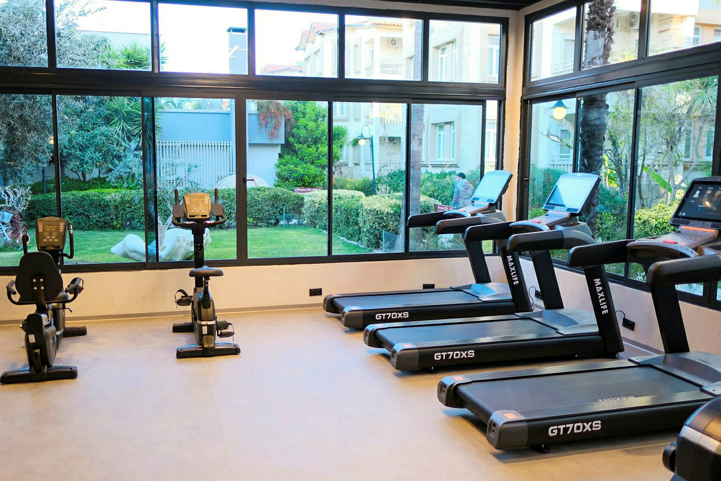 Ett gym med löpband och motionscyklar framför fönster med utsikt mot en trädgård. (AI-genererad text)