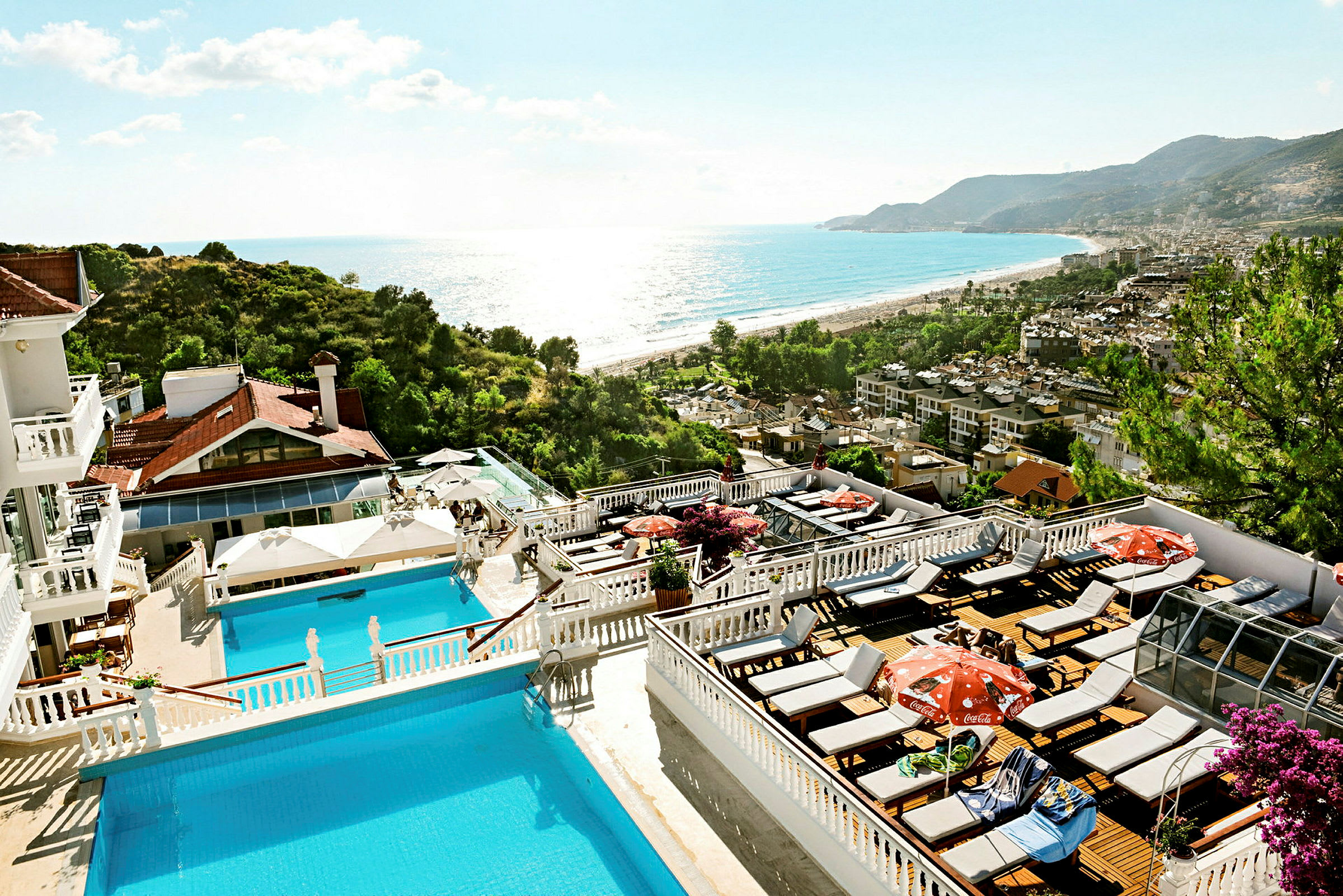 Bild av ett hotellområde med pooler och solterrasser, omgivet av grönska och med utsikt över havet och en strand. (AI-genererad text)