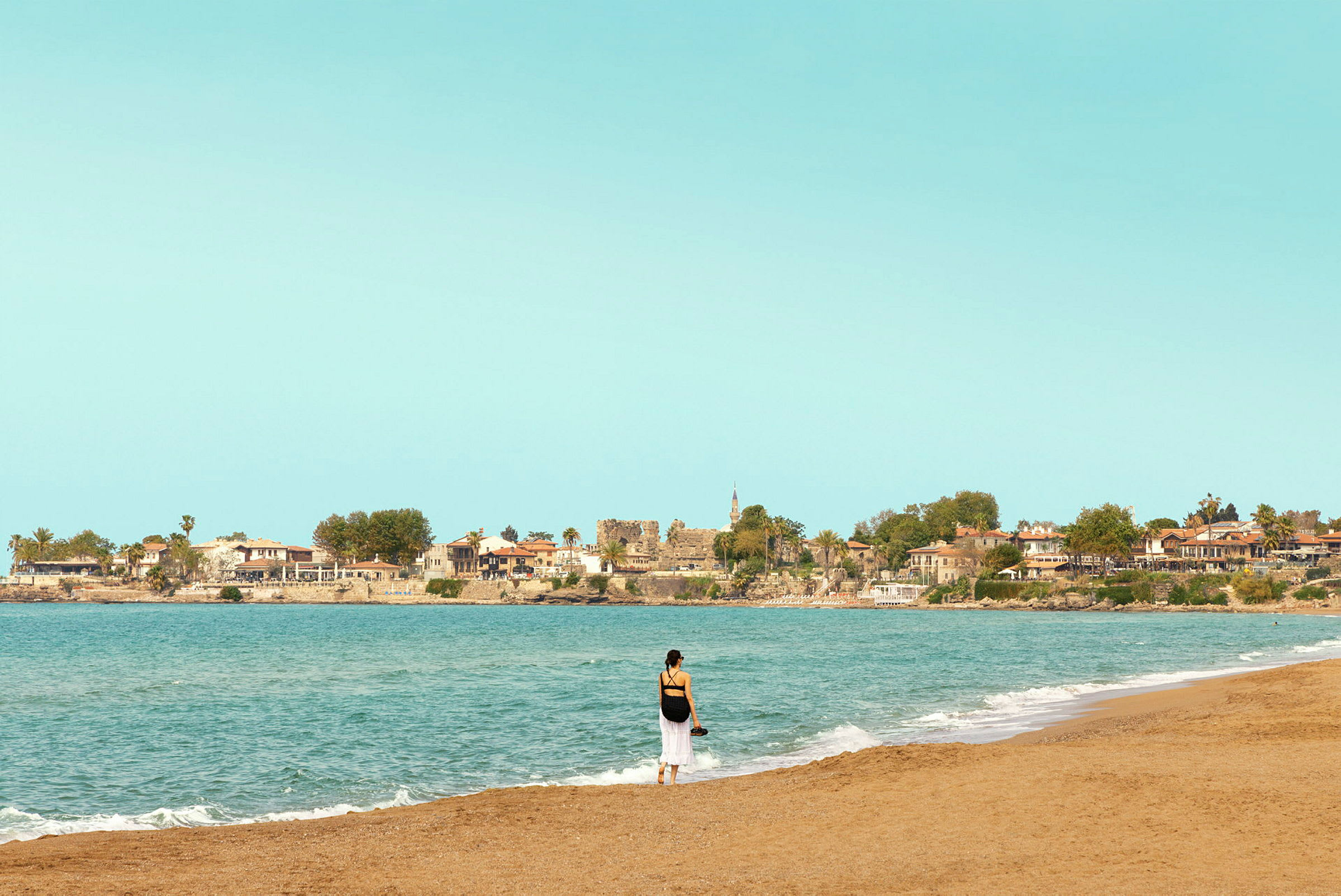 En person står vid strandkanten med hav och en liten stad i bakgrunden. (AI-genererad text)